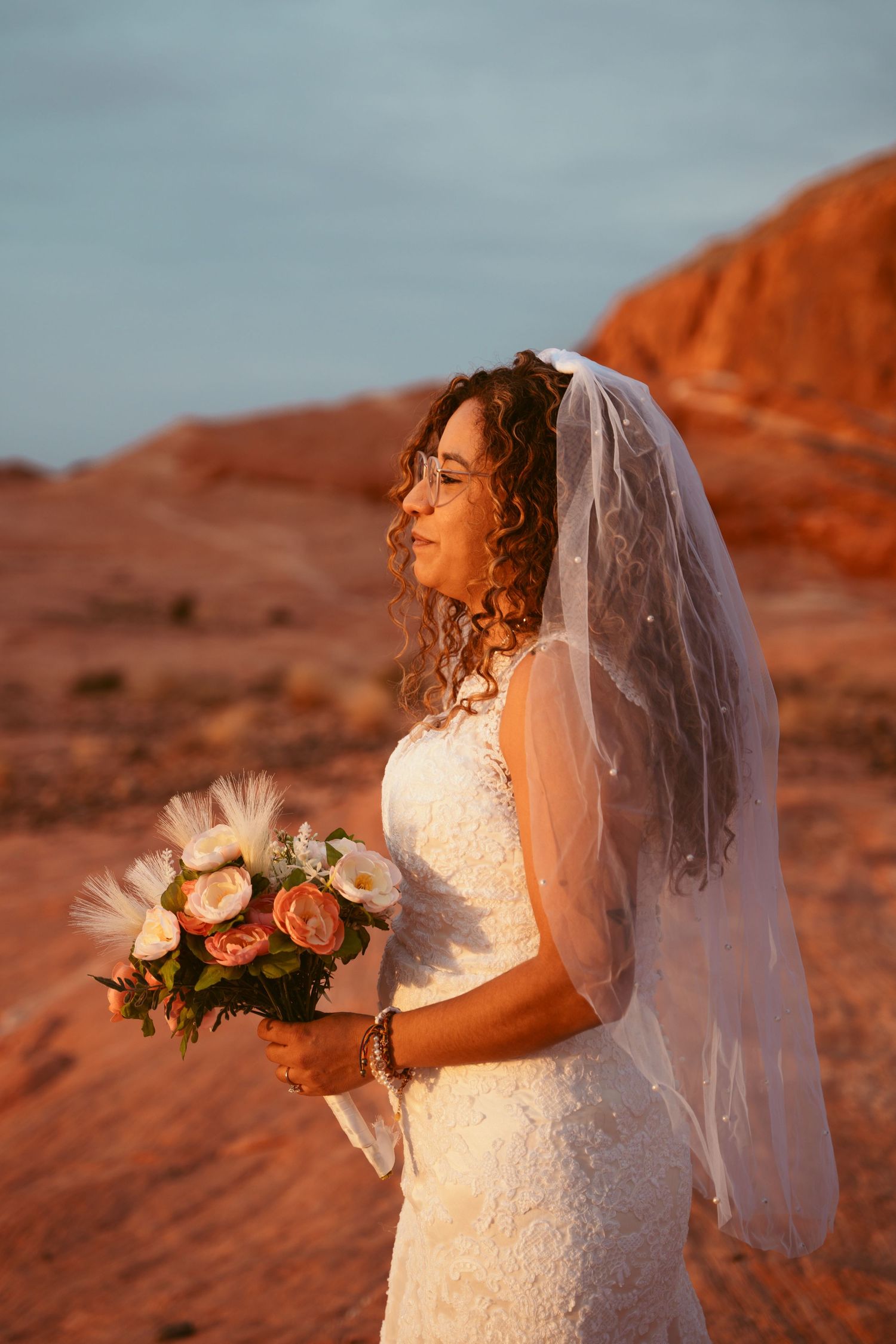 Bride in white dress holds bouquet against dramatic red desert landscape at sunset.