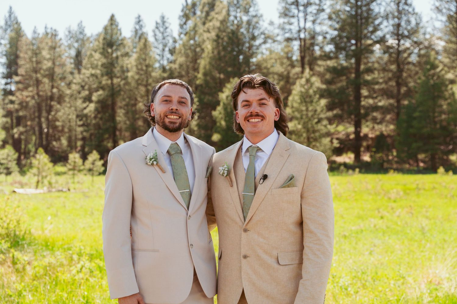 Groomsmen in khaki suits share a laugh while standing in a grassy field with evergreen forest backdrop.