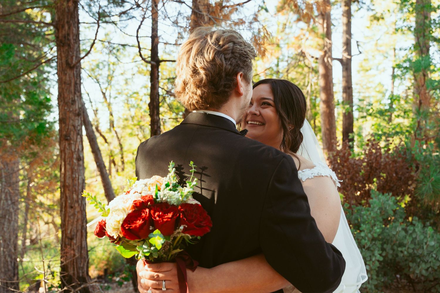 A joyful wedding embrace in a sun-dappled forest with red flowers.