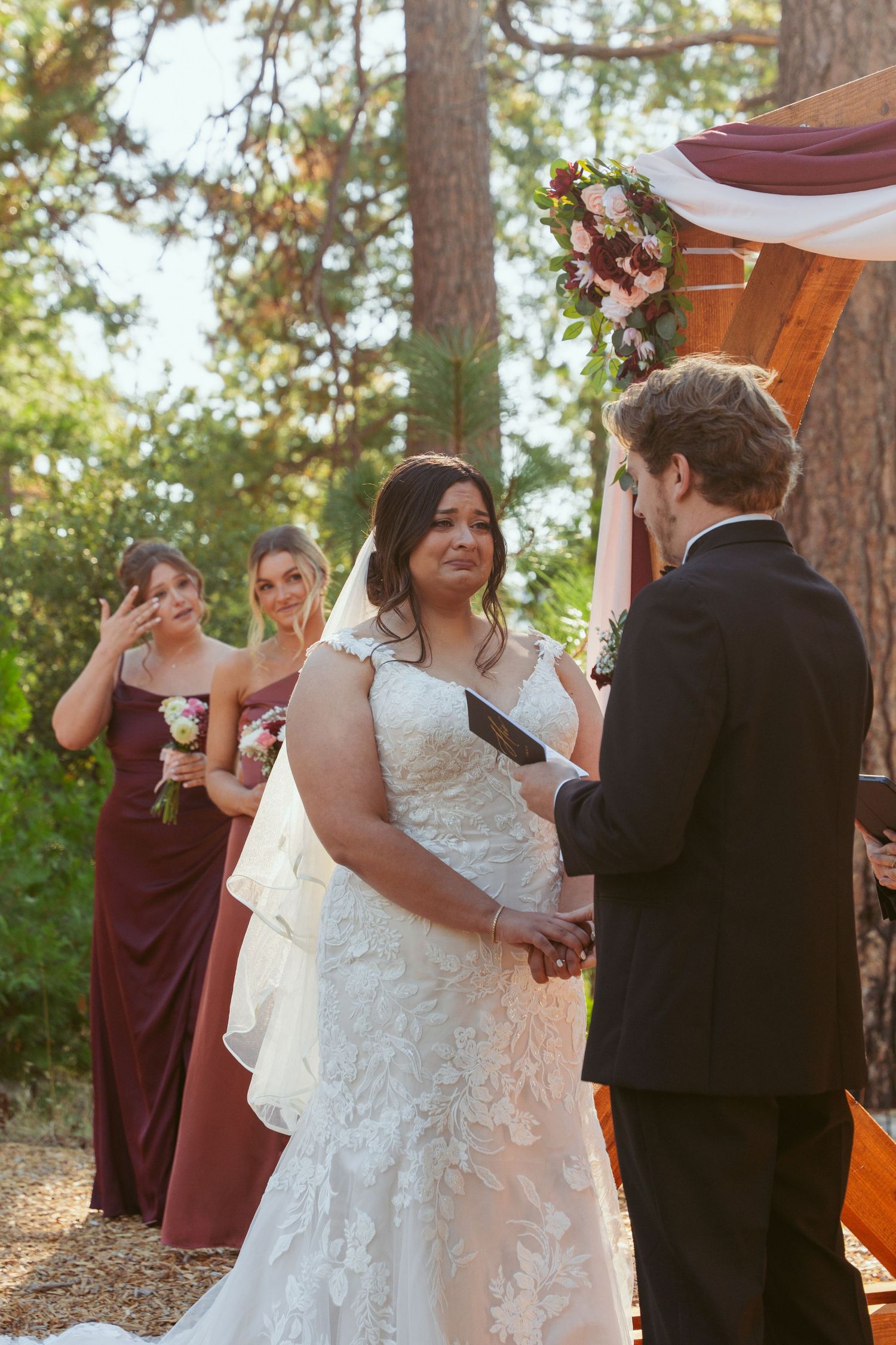 Wedding ceremony moment captured in a sunlit forest setting with burgundy and white decor.