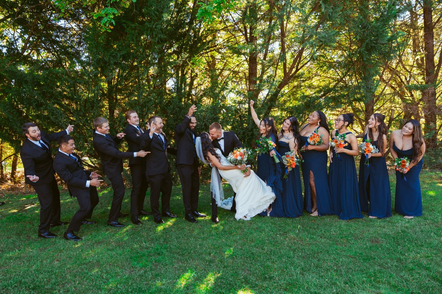 Wedding party strikes fun poses on grassy lawn with bridesmaids in blue and groomsmen in black.