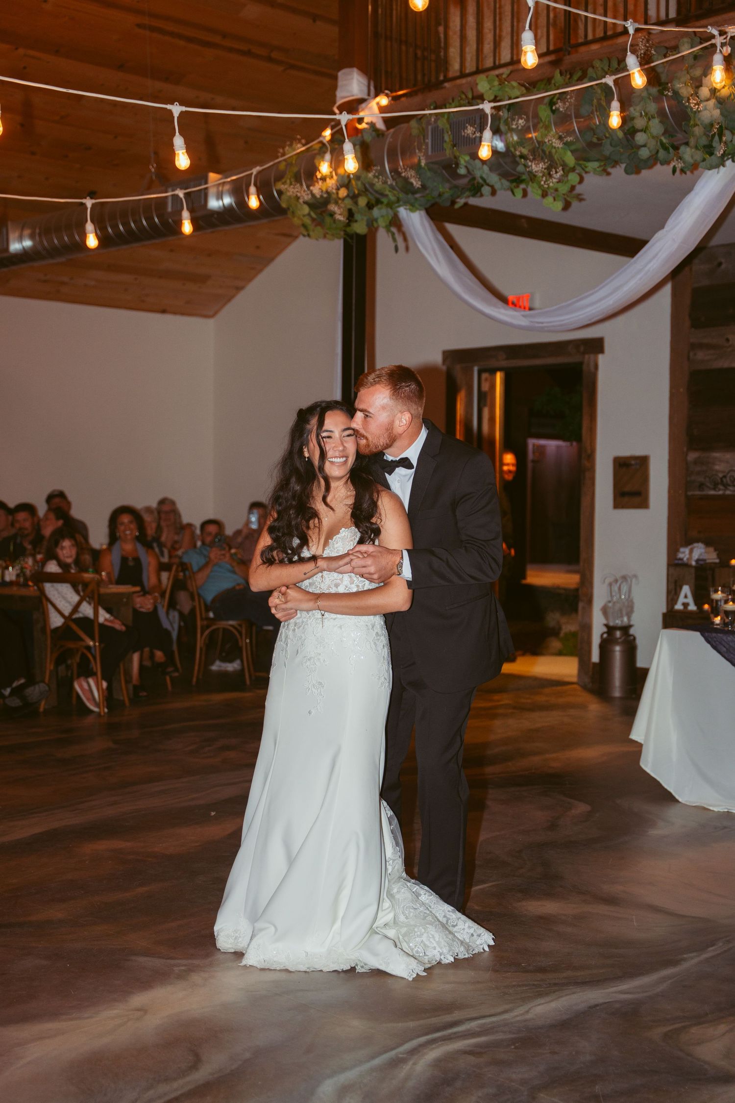 Newlyweds share first dance under string lights in rustic barn reception venue.