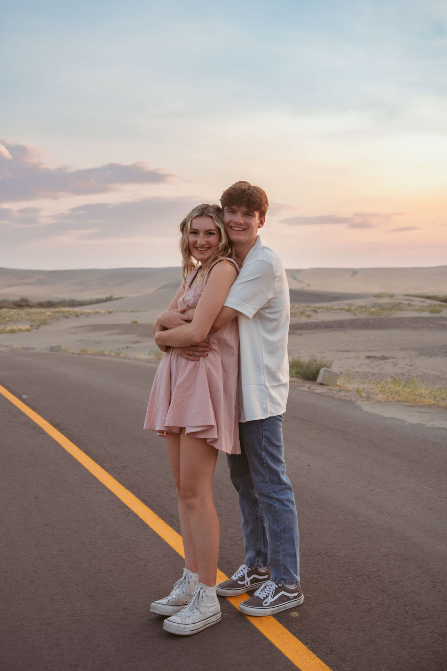 A couple in casual attire stands together playfully on a desert highway at dusk.
