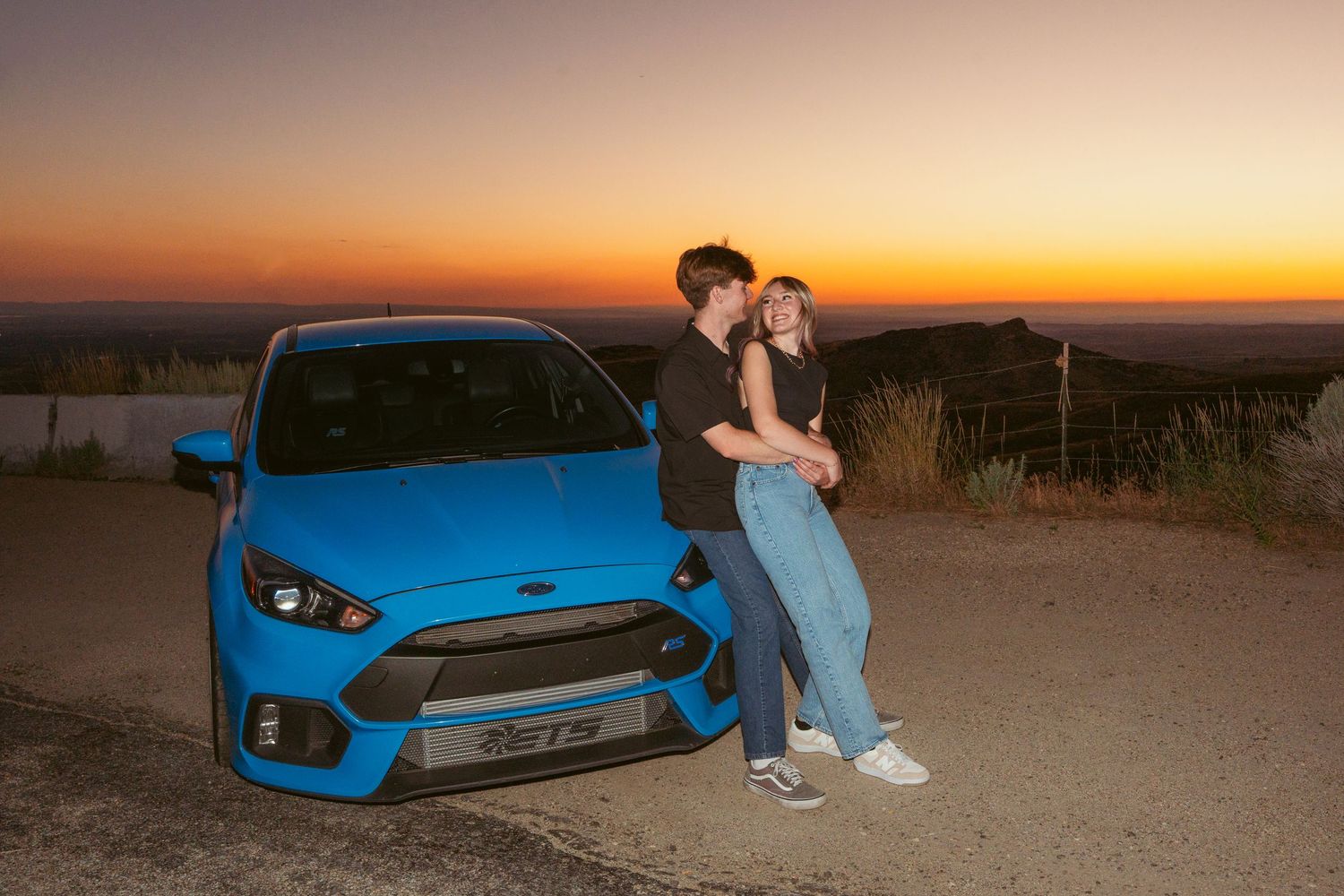 Couple leaning against blue Ford Focus RS at sunset overlook.