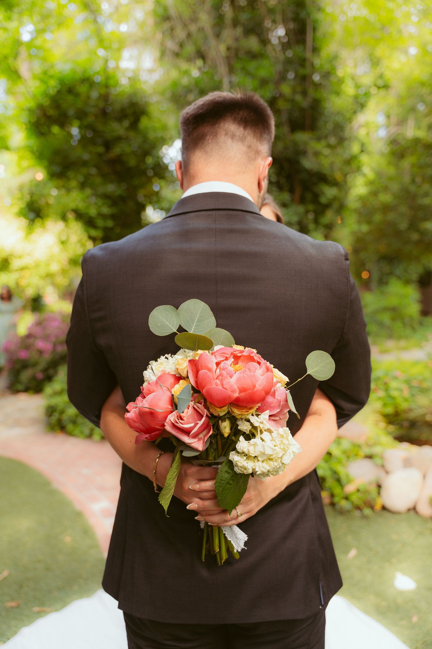 Groom in dark suit holding coral peony bouquet behind his back in garden setting.