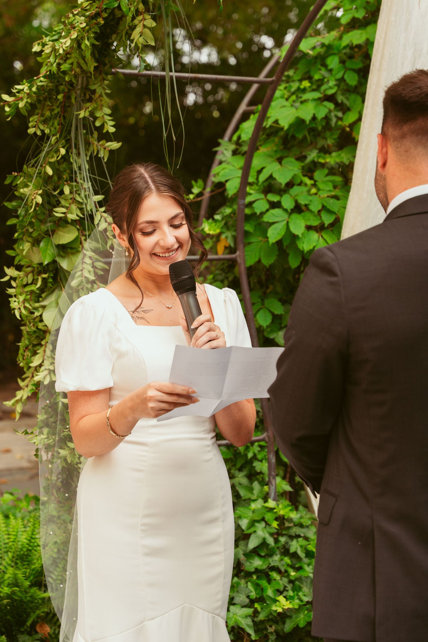 A bride in a white dress reads from paper during an outdoor garden wedding ceremony.