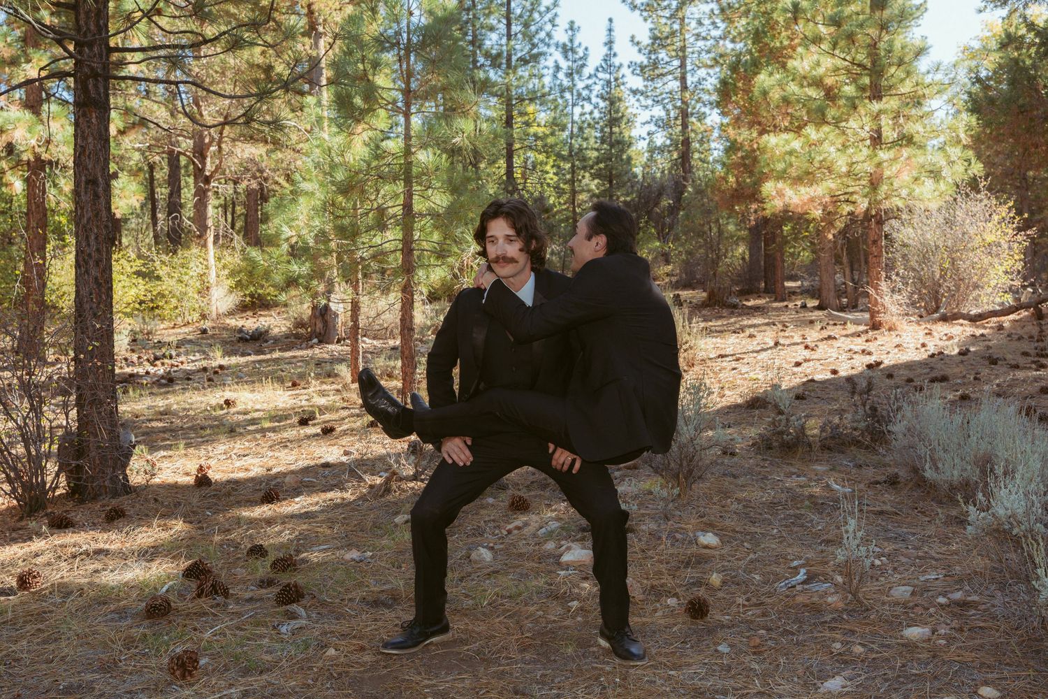 Two people in dark clothing strike playful poses in a forest setting.