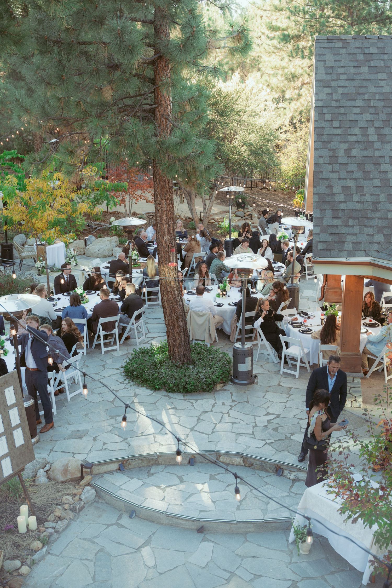 Outdoor wedding reception setup with white chairs and stone patio surrounded by pine trees.
