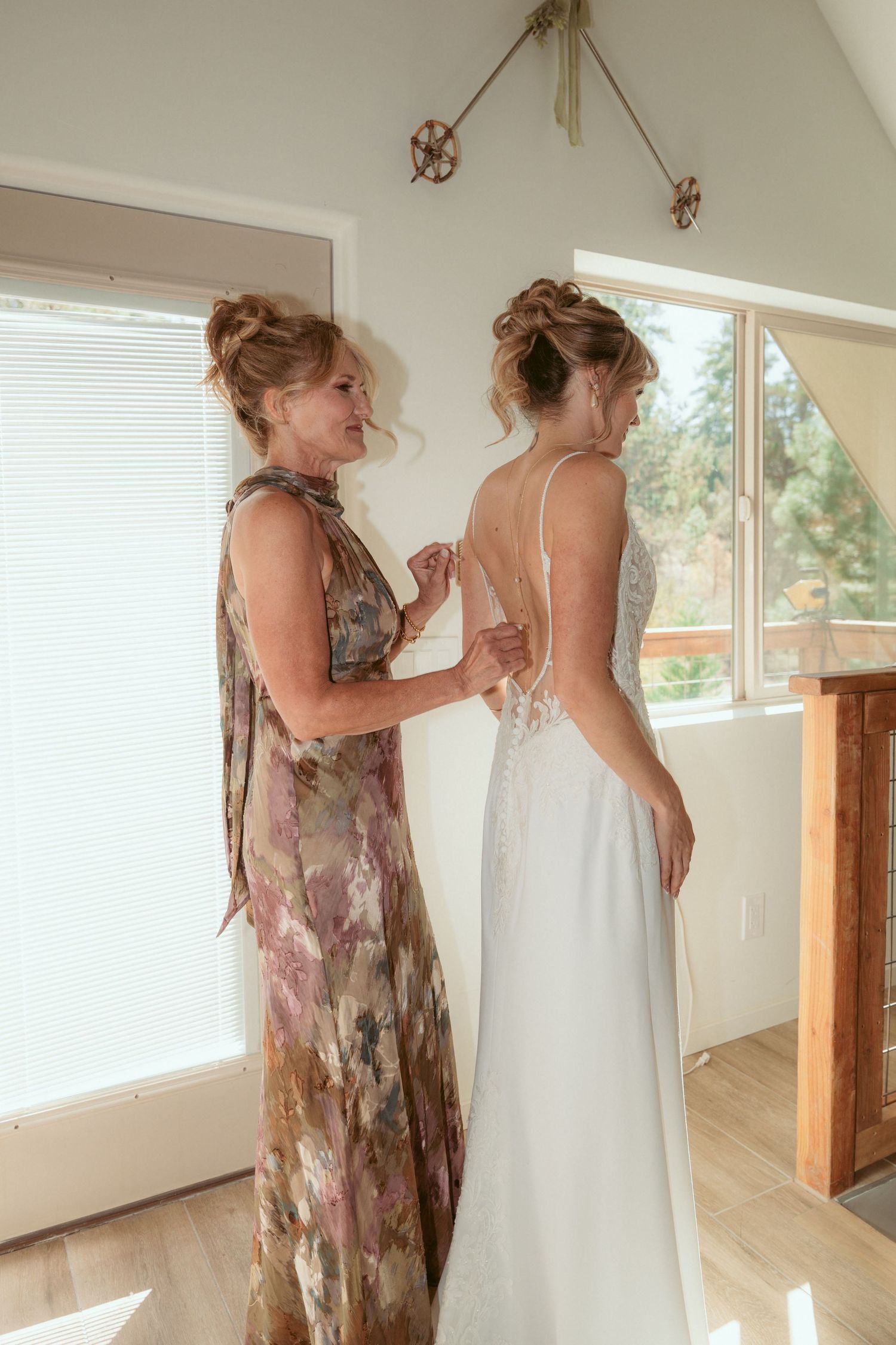 Two people in a bright room, one wearing a floral dress helping another with a white wedding dress.