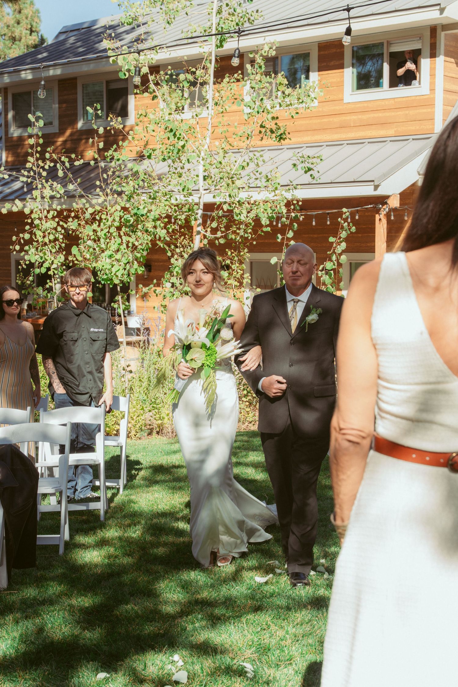 Wedding ceremony procession taking place in a sunlit garden with white chairs.