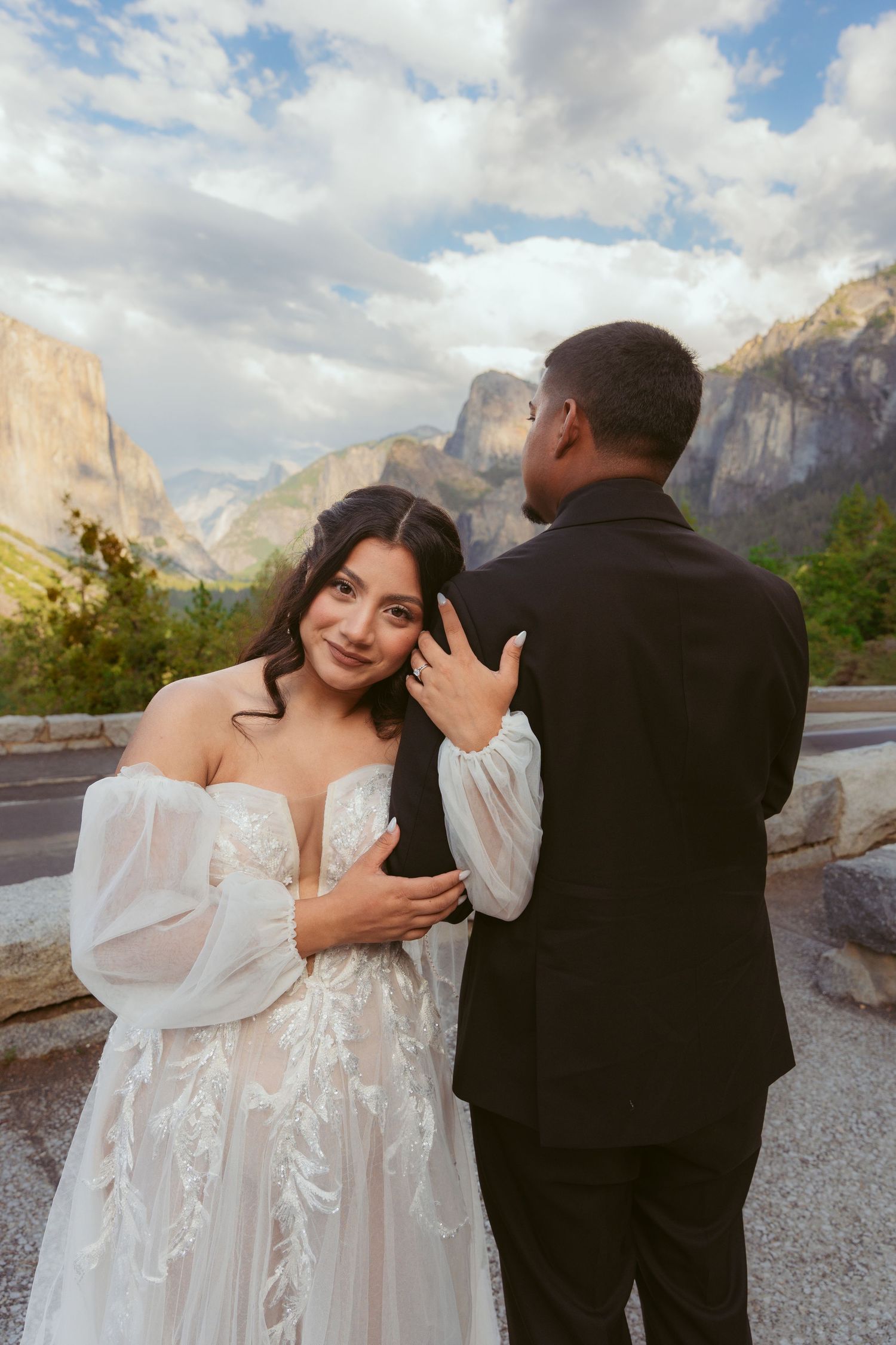 Bride in flowing white dress poses with groom against scenic mountain landscape.