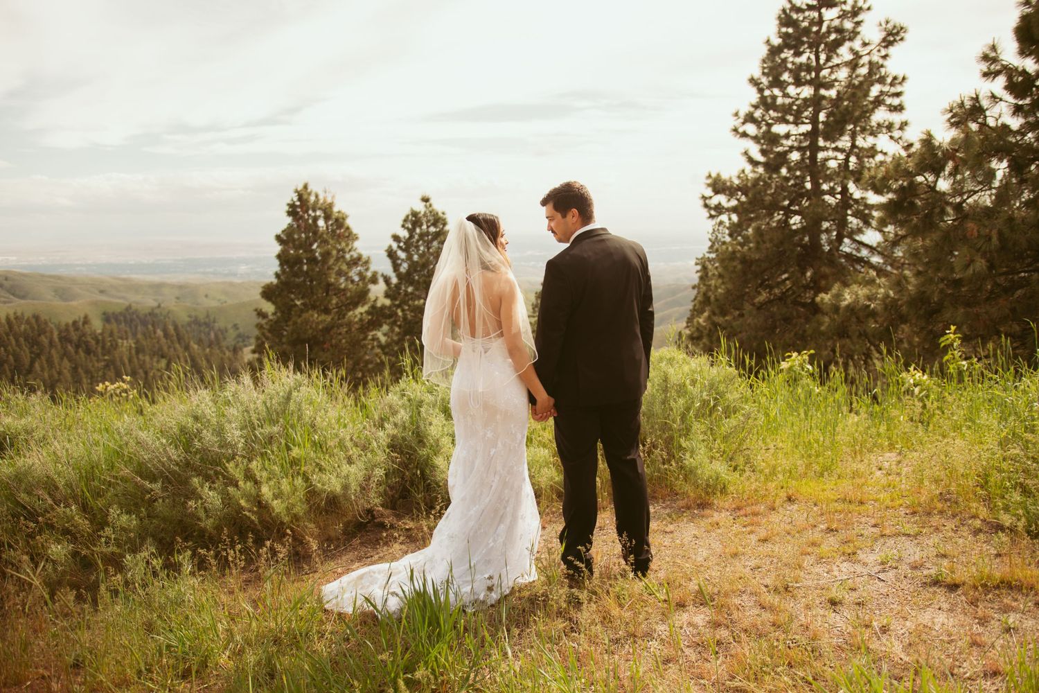 A wedding couple stands on a mountain trail overlooking a scenic valley landscape.