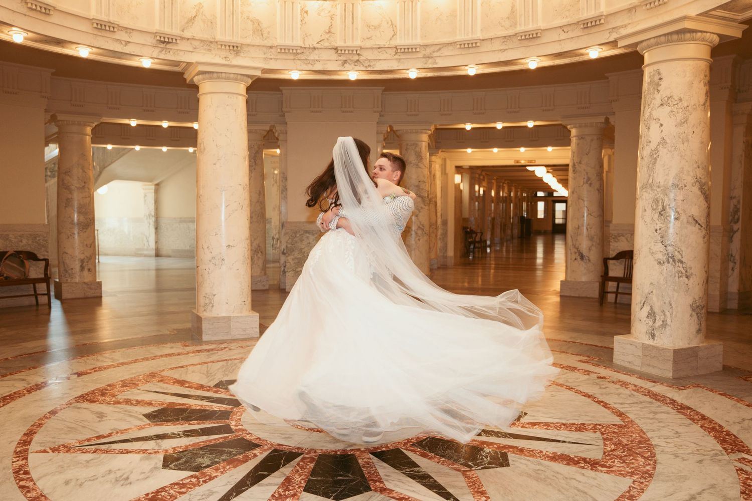 Bride twirling in white dress inside ornate rotunda with marble floors.
