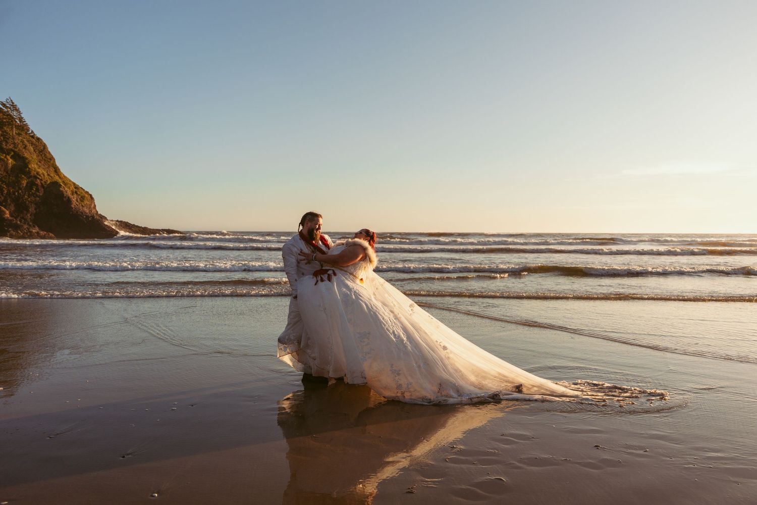 Couple in wedding attire walking along the ocean shore at sunset.