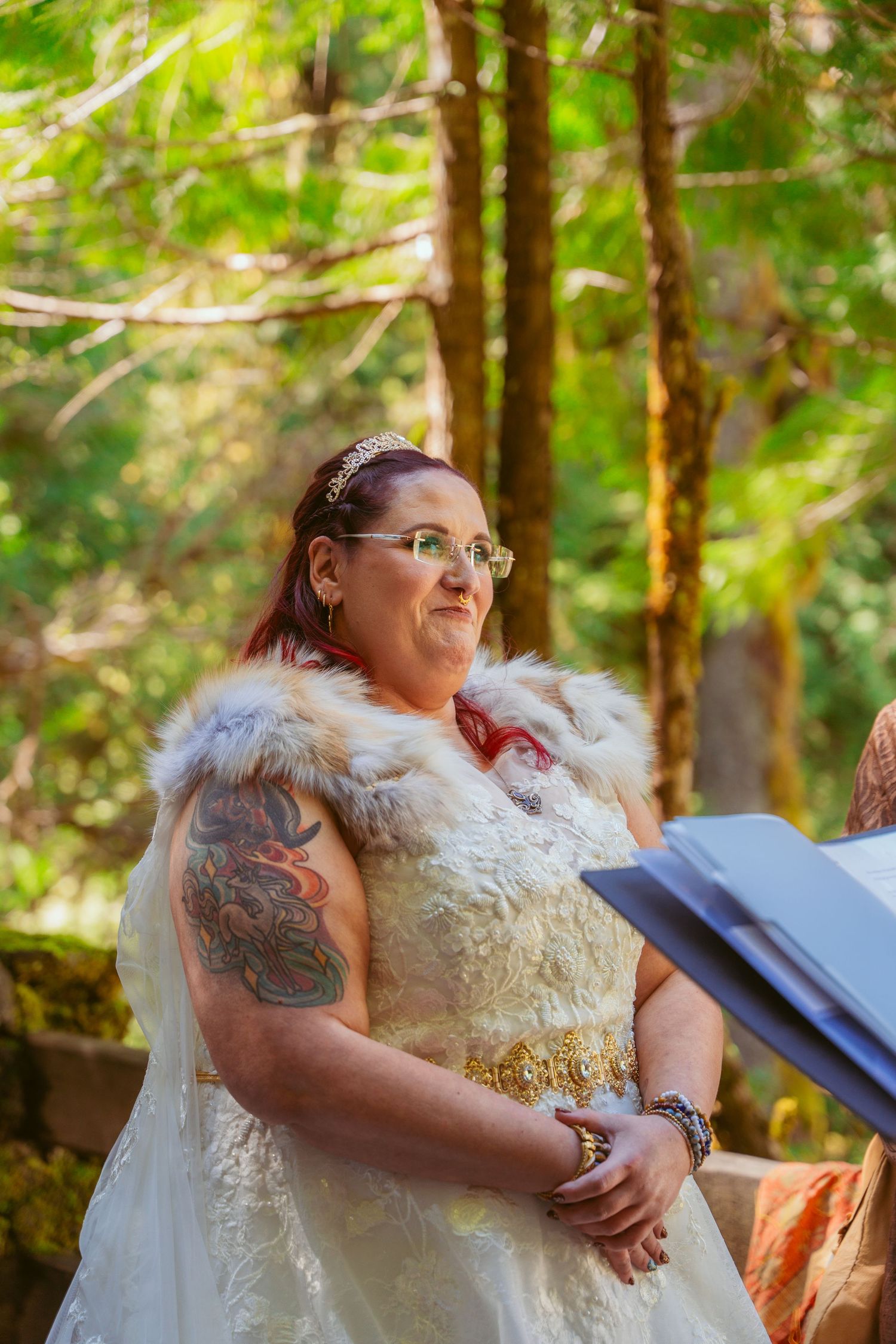 A bride in a white gown with fur trim stands in a sunlit forest setting during an outdoor ceremony.