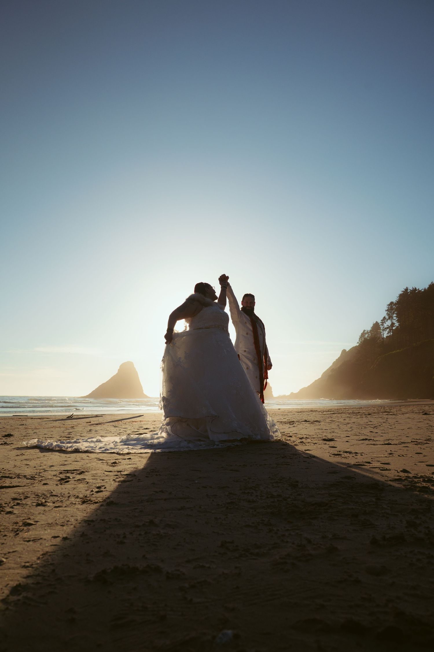 A wedding couple's silhouette against the sunset on a beach with coastal rock formations.