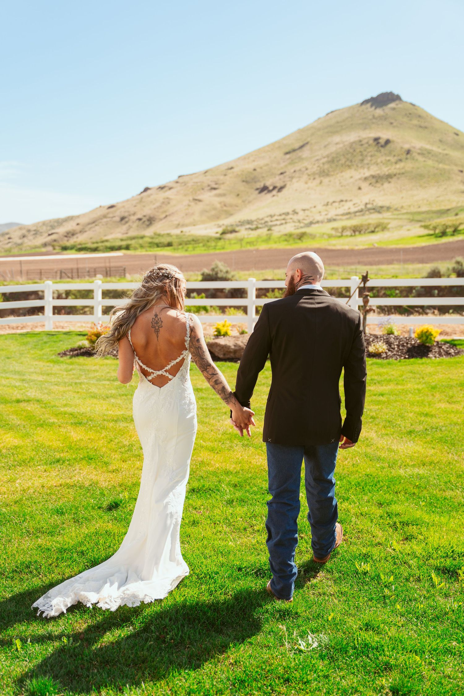 Couple walks away from camera across green field with mountains and white fence visible.