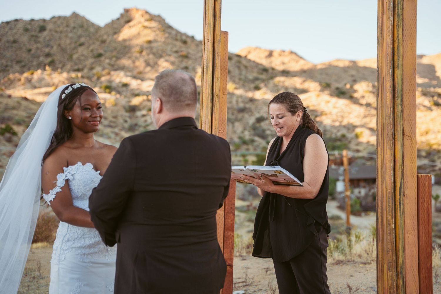 An intimate desert wedding ceremony with an officiant and couple under a rustic wooden arch at sunset.