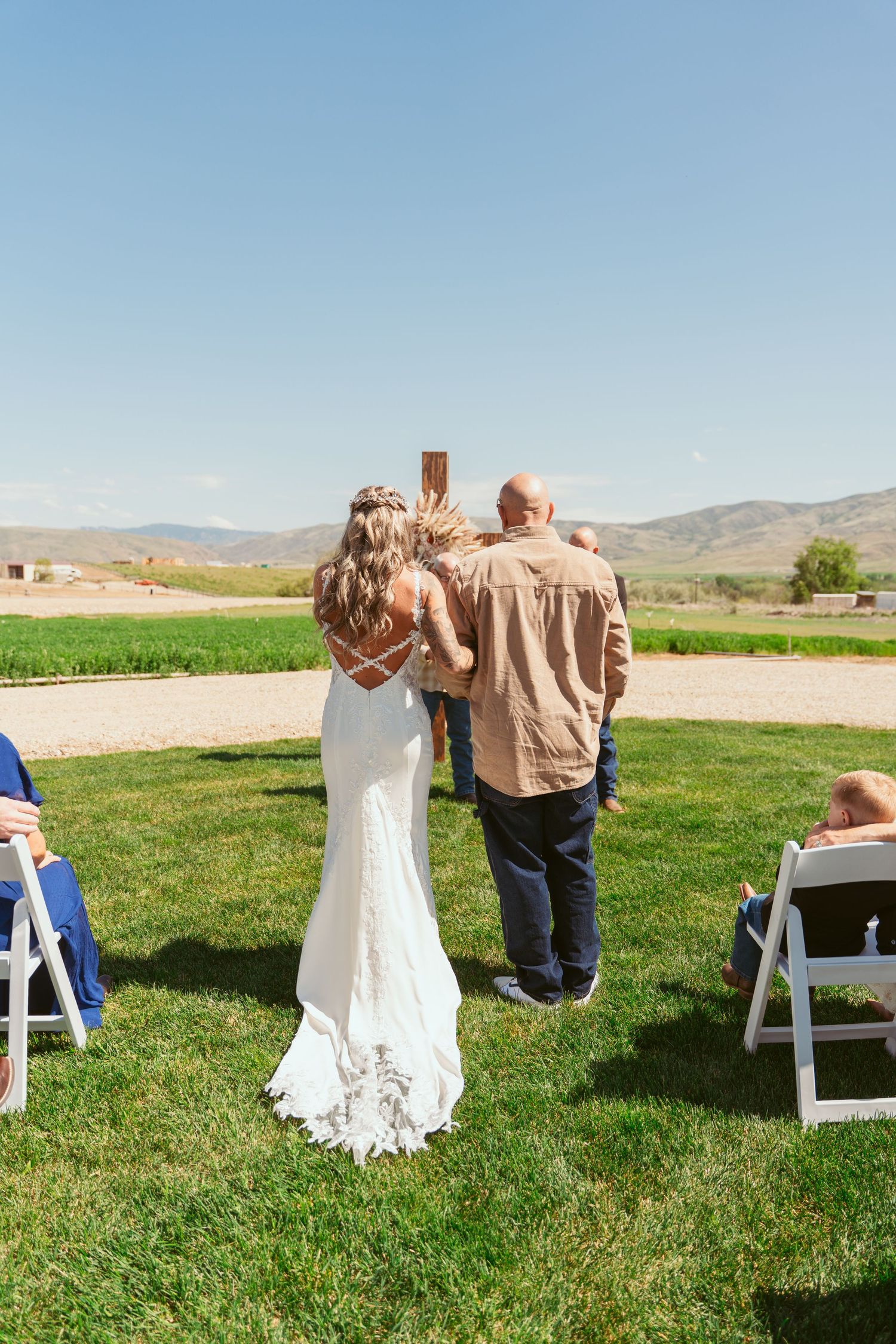 Intimate outdoor wedding ceremony with white chairs and wooden cross on a sunny spring day.