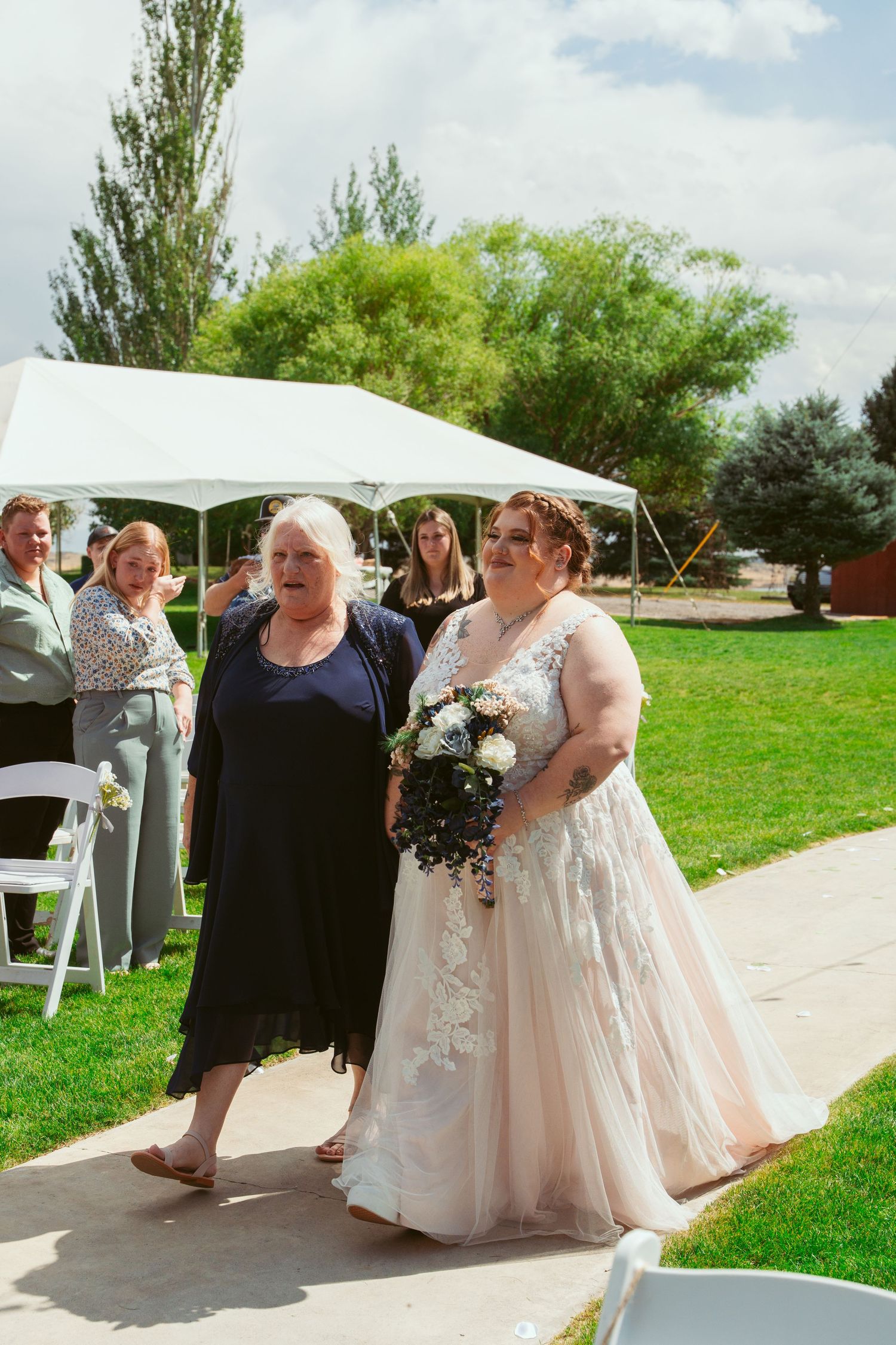 Wedding attendees in formal attire gather on a paved pathway surrounded by green grass and trees.