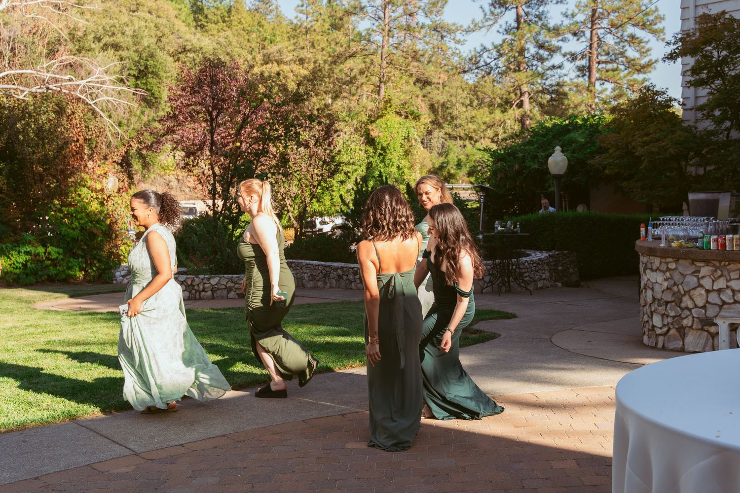 Bridesmaids in elegant green dresses walking on outdoor stone patio during wedding celebration.