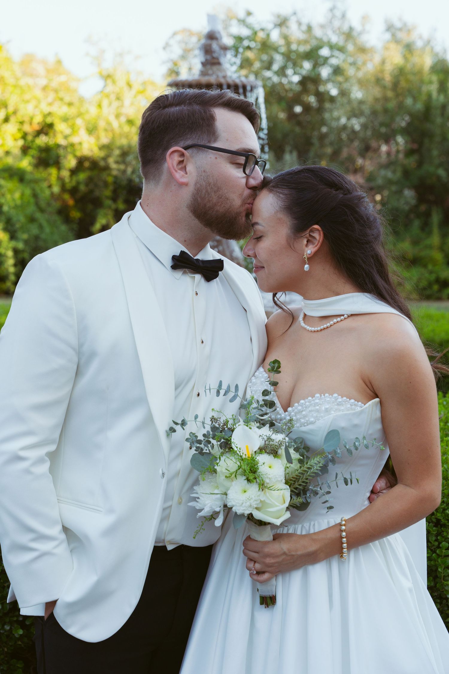 A couple in white formal wear embraces while holding a delicate bouquet of white flowers.
