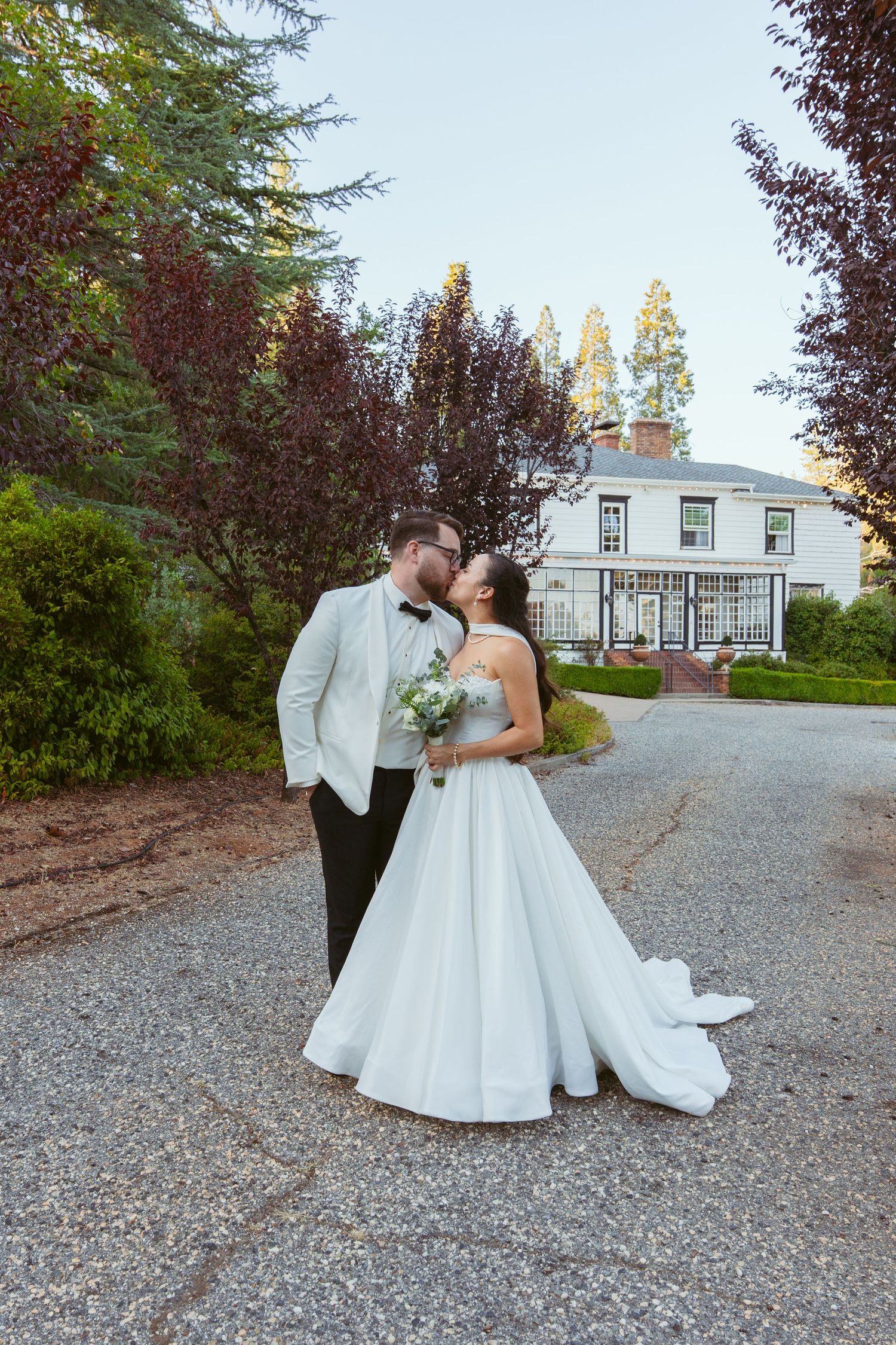 Wedding couple posing on a gravel path with a white building visible in the background.