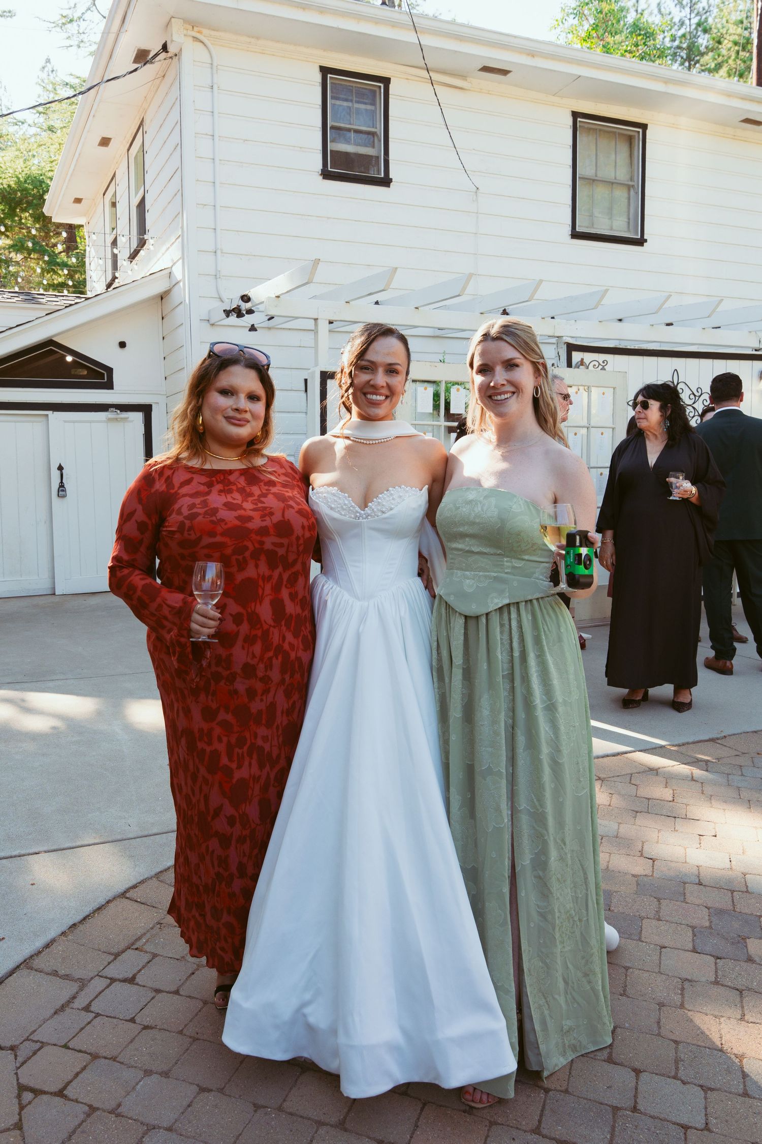 Three women pose together at a wedding, with the bride in white between two guests in formal attire.