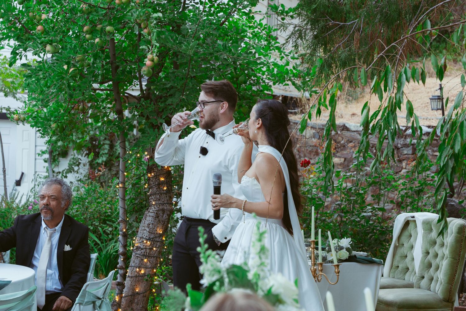 Wedding celebration under green trees with elegant outdoor seating arrangements.