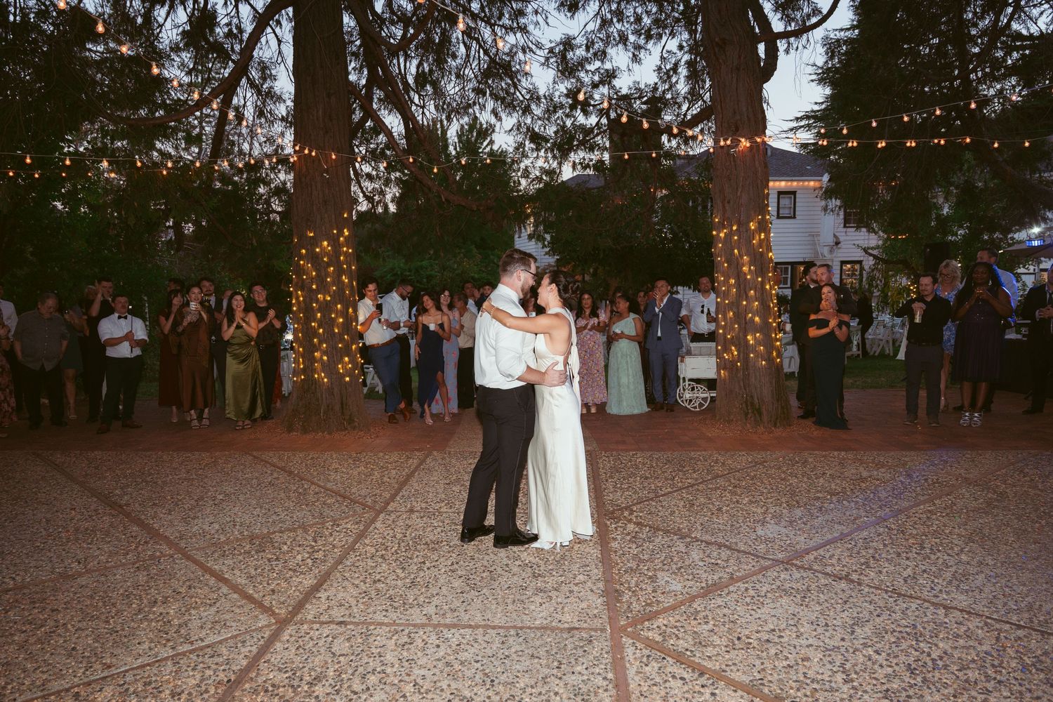 Evening wedding celebration with guests dancing under illuminated trees and string lights.