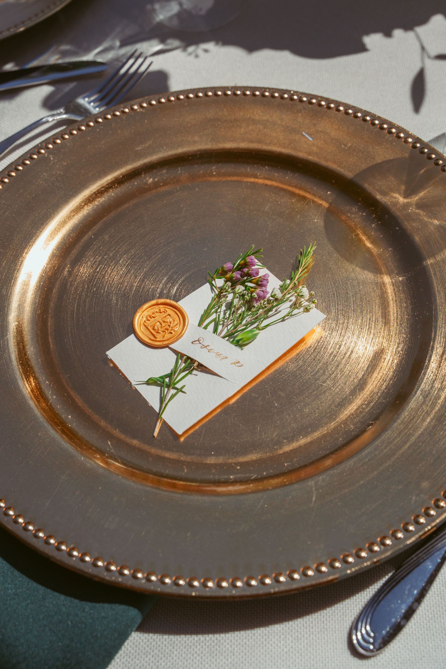 Gold charger plate with a wax-sealed place card and small floral sprig.