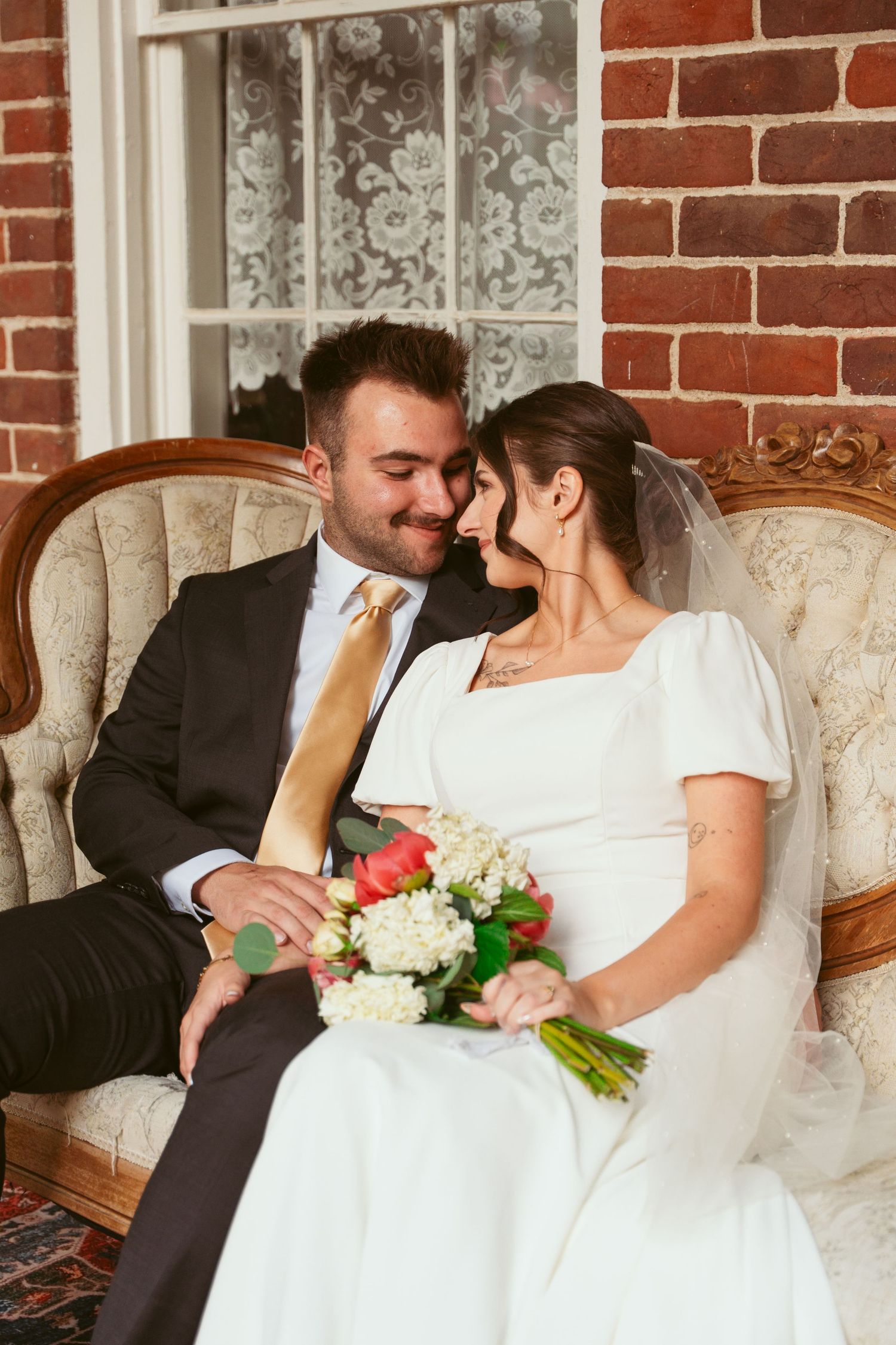 A romantic wedding scene showing a couple seated on an antique sofa against a brick wall with white lace curtains.