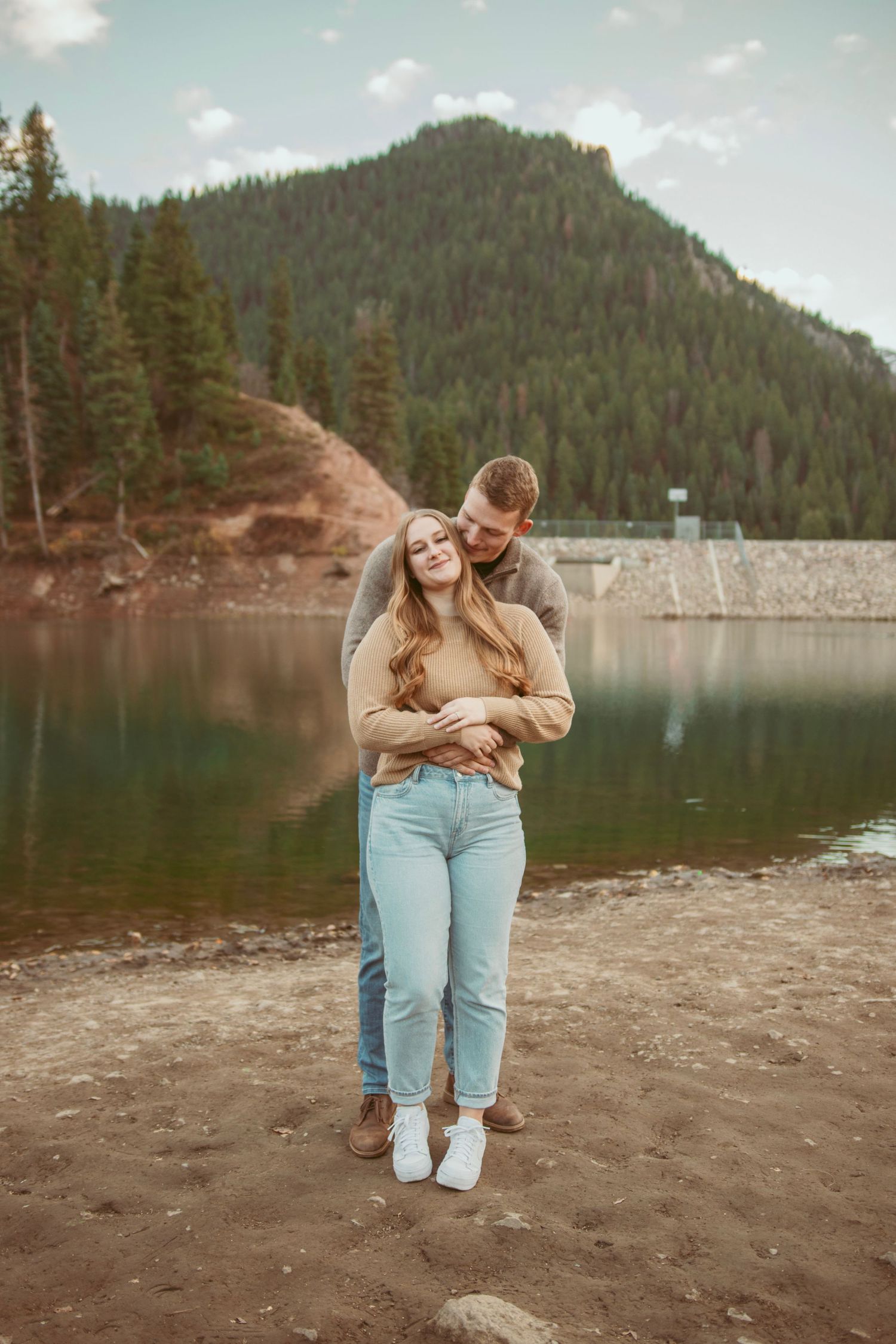 Romantic photoshoot at a lakeside beach with mountains and forest in the background.