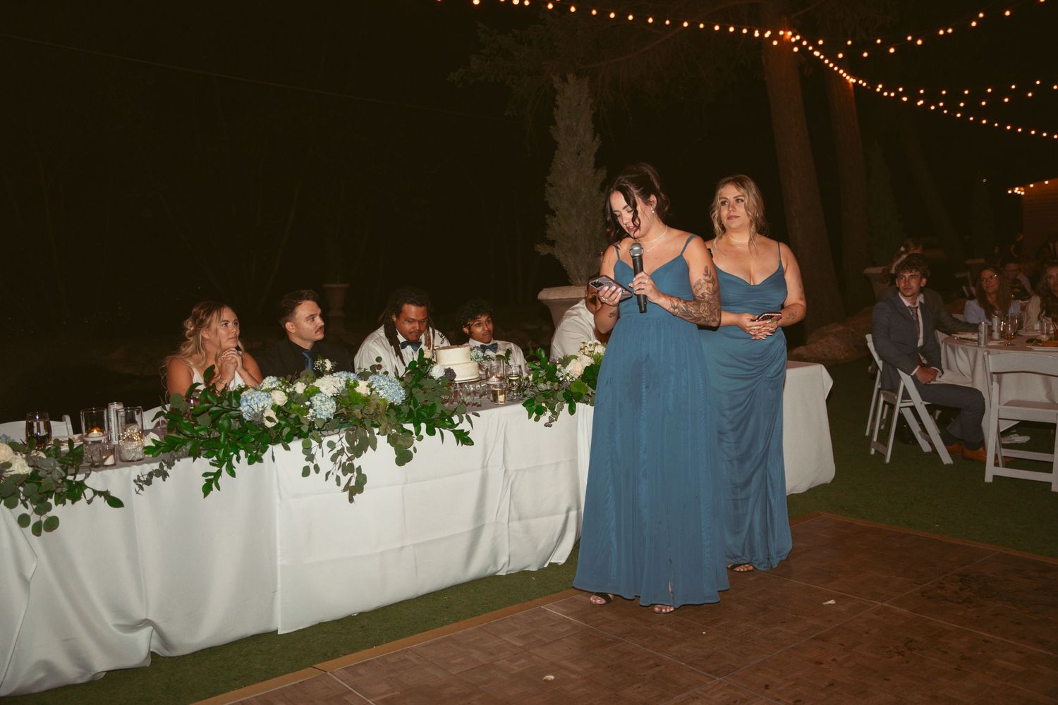 Wedding guests dancing together on a wooden dance floor under string lights at an outdoor evening reception.
