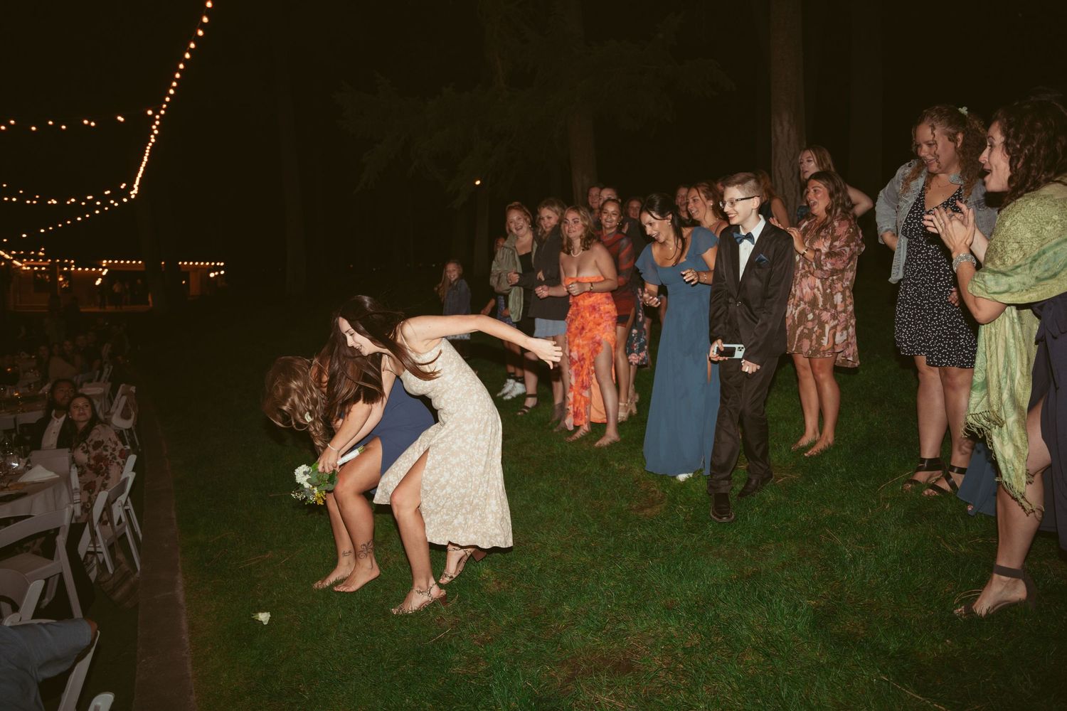 Wedding guests dance and celebrate on a grassy lawn under string lights at night.