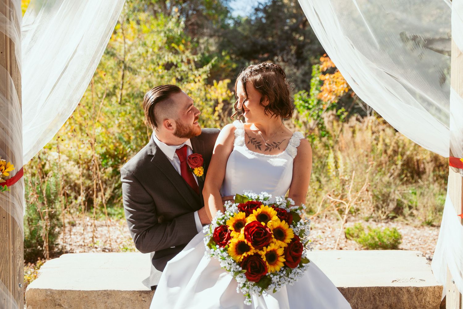 Wedding couple poses with vibrant sunflower and rose bouquet under draped outdoor pavilion.