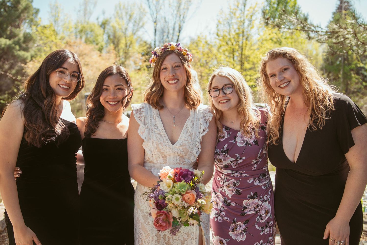 A group of bridesmaids in black and floral dresses pose together outdoors.