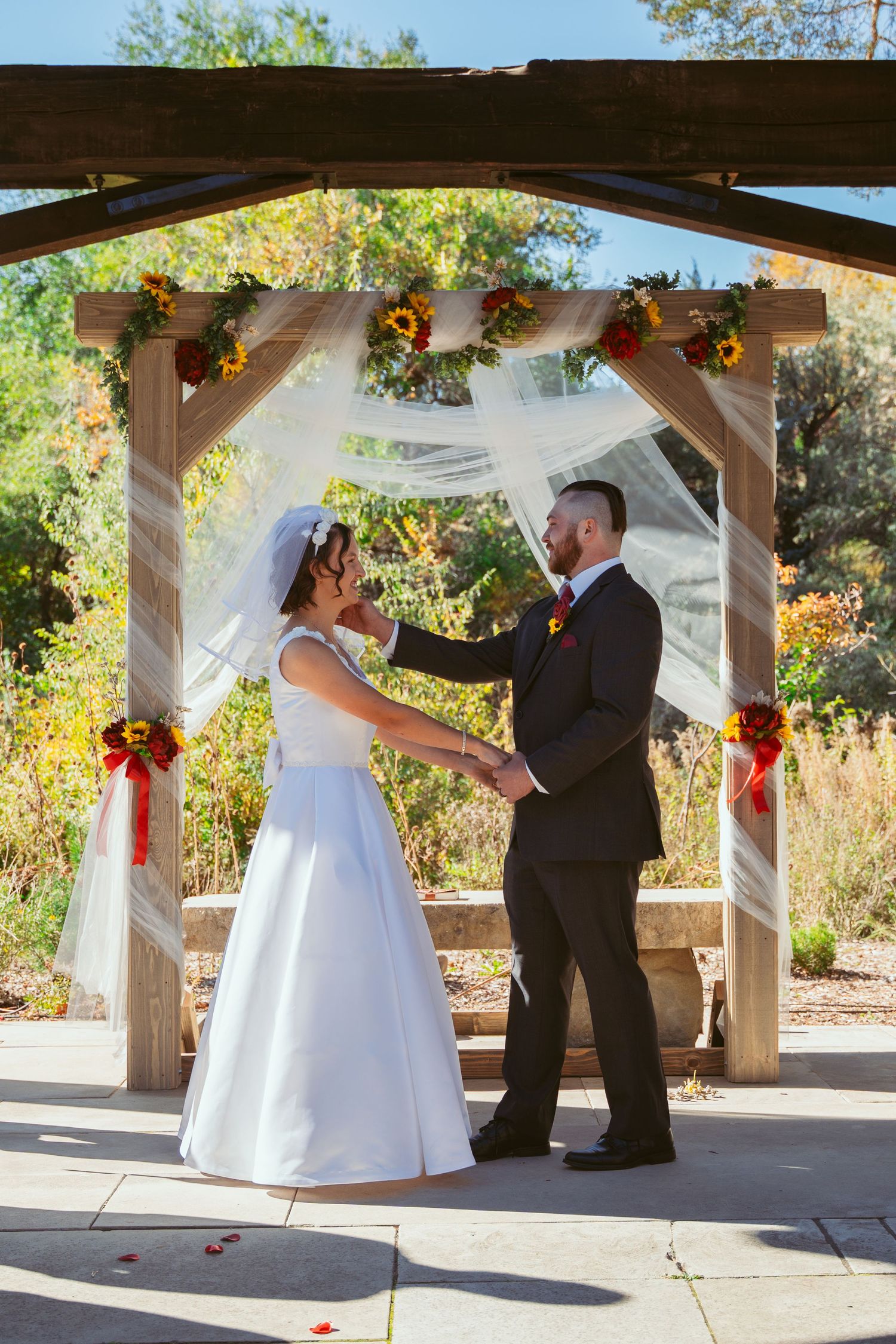 Wedding ceremony under a wooden pergola decorated with red bows and white fabric.