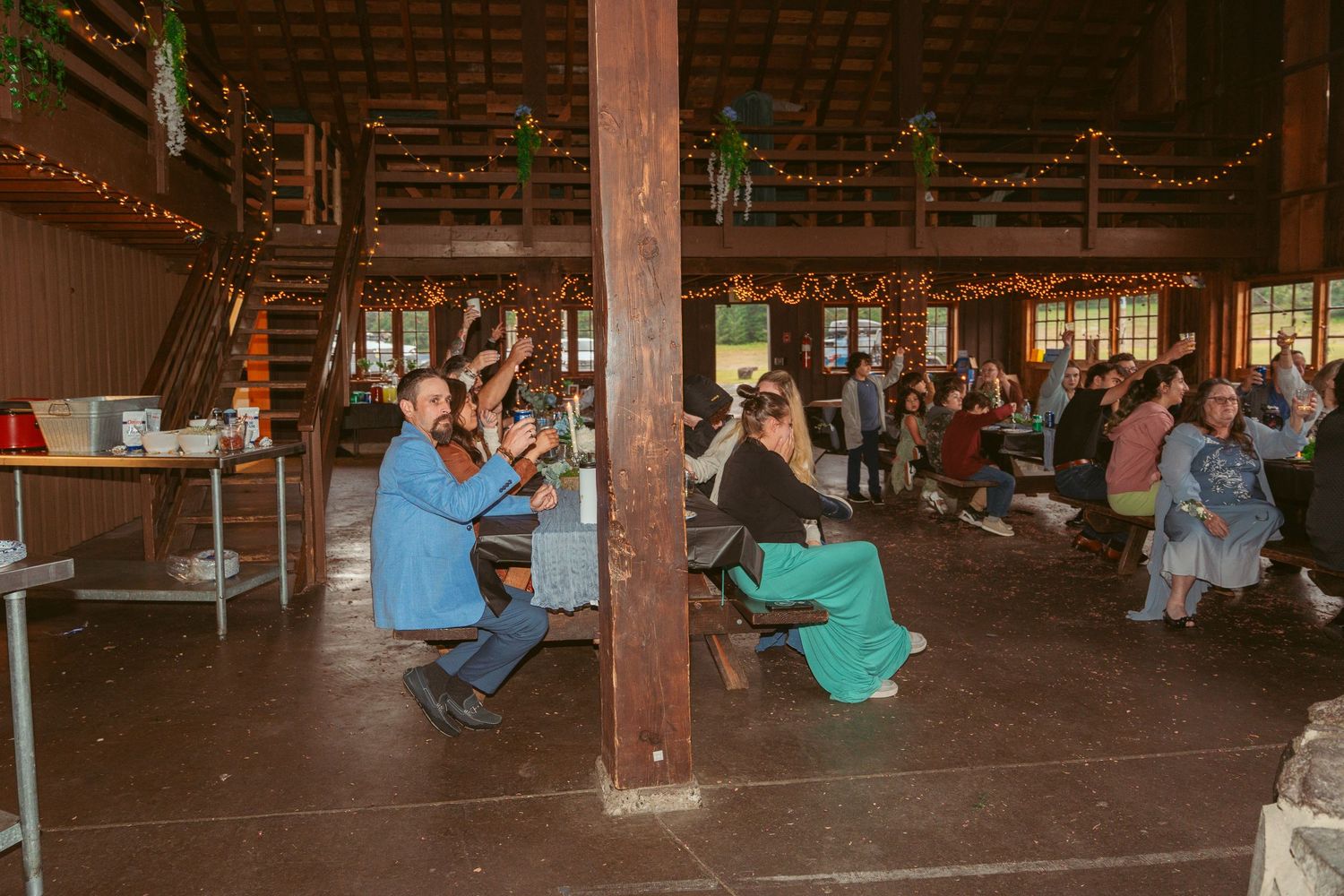 Wedding guests seated in wooden barn venue with string lights overhead during reception.