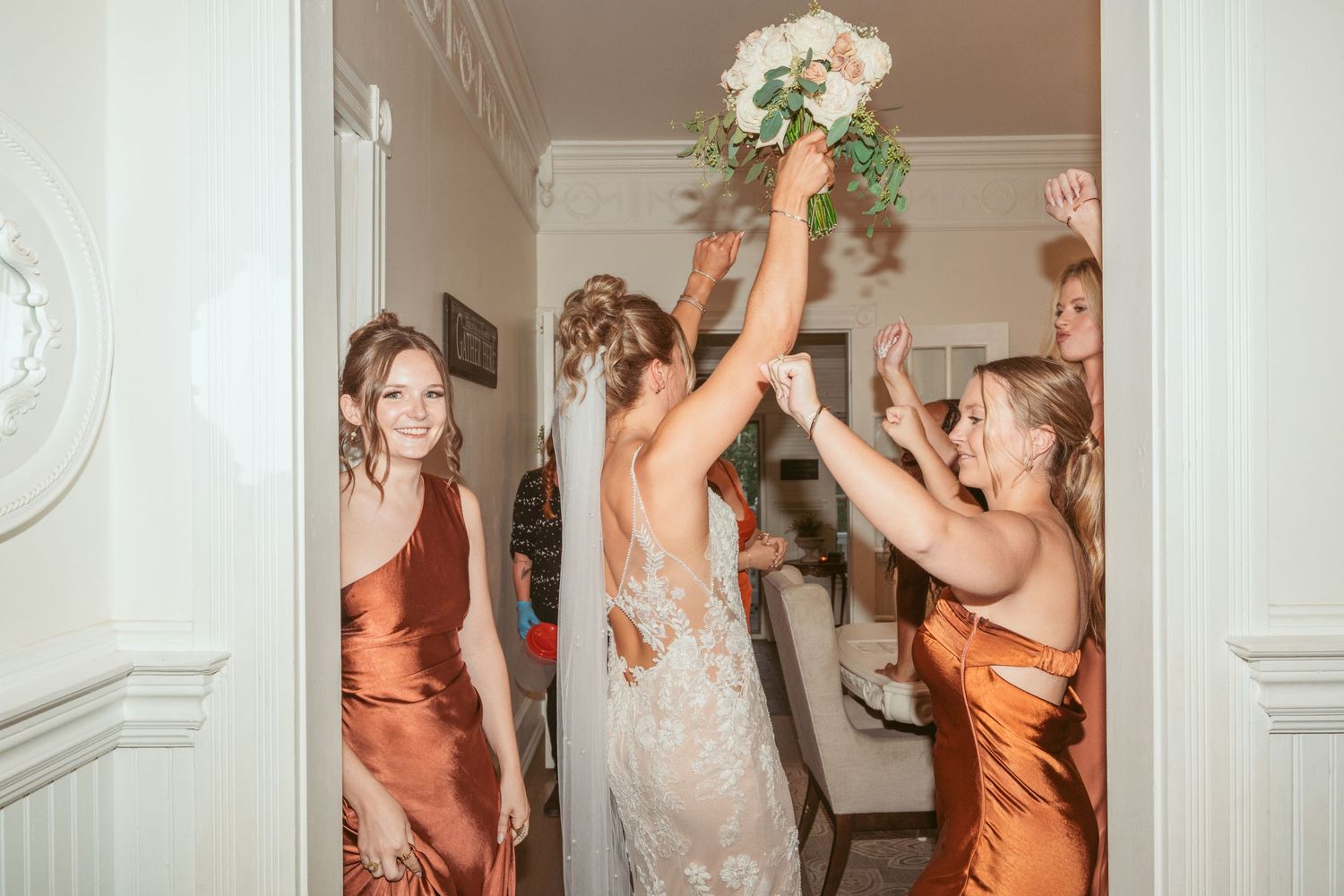 Wedding party celebrates with energetic dancing in an elegant white-walled indoor space.
