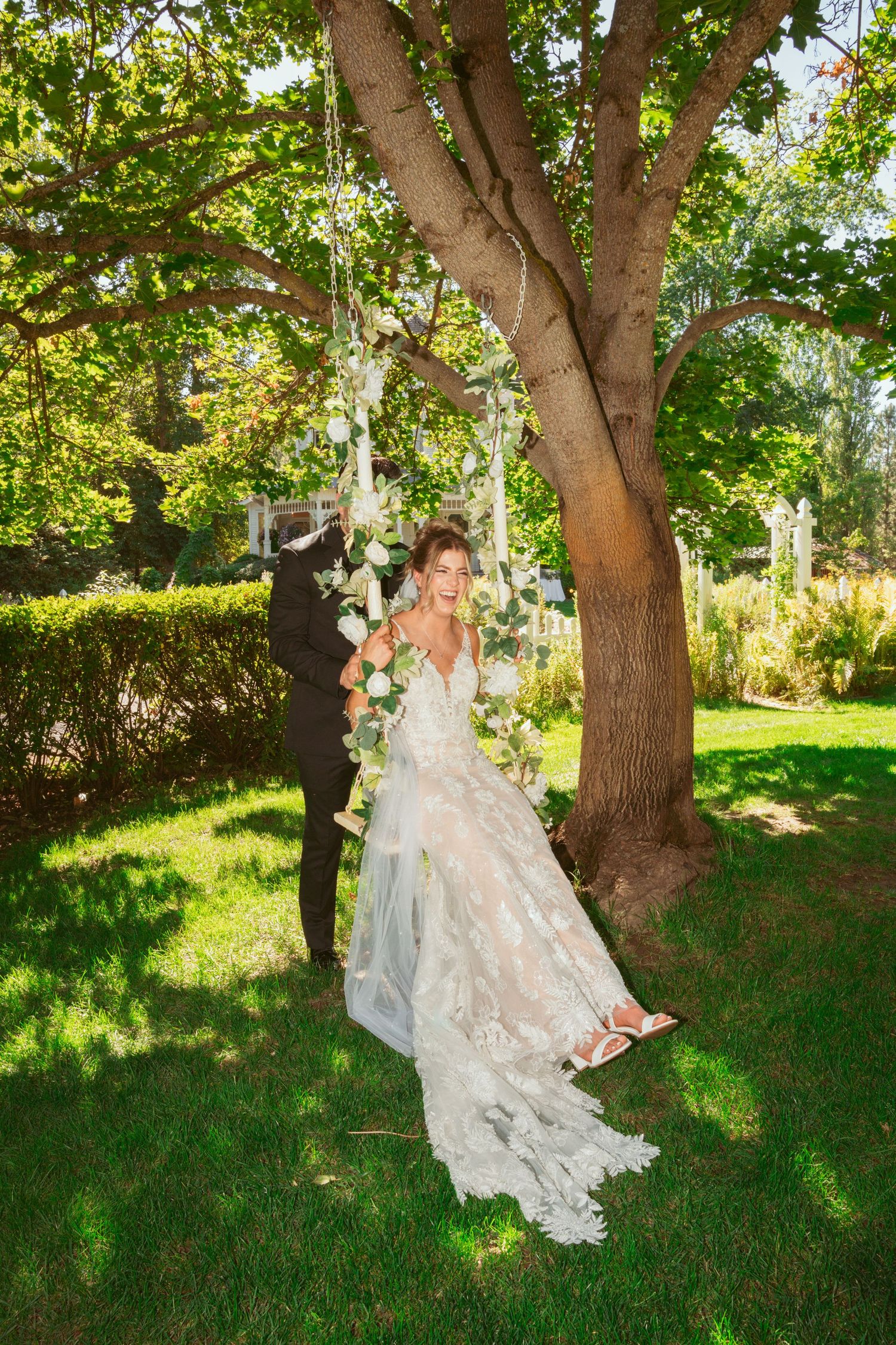 Bride in lace gown poses on a floral swing hanging from a large tree in a garden setting.