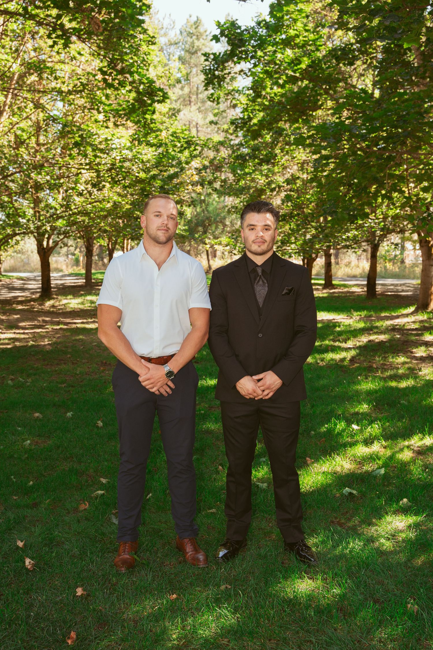 Two people in formal attire stand side by side on green grass in a sunny orchard setting.