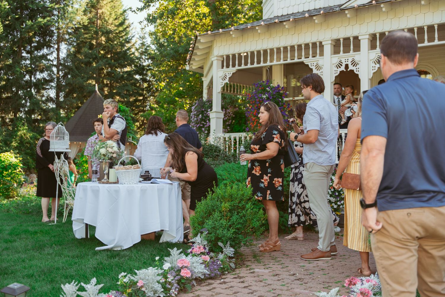 Wedding guests line up at an outdoor reception buffet table near a white gazebo surrounded by gardens.