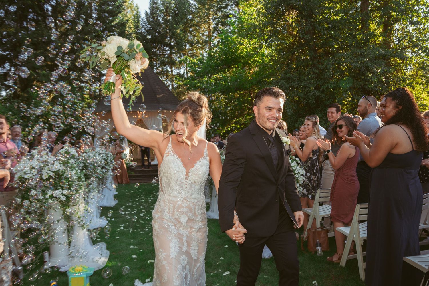 Newlyweds celebrate while walking down the aisle as guests cheer in an outdoor garden setting.