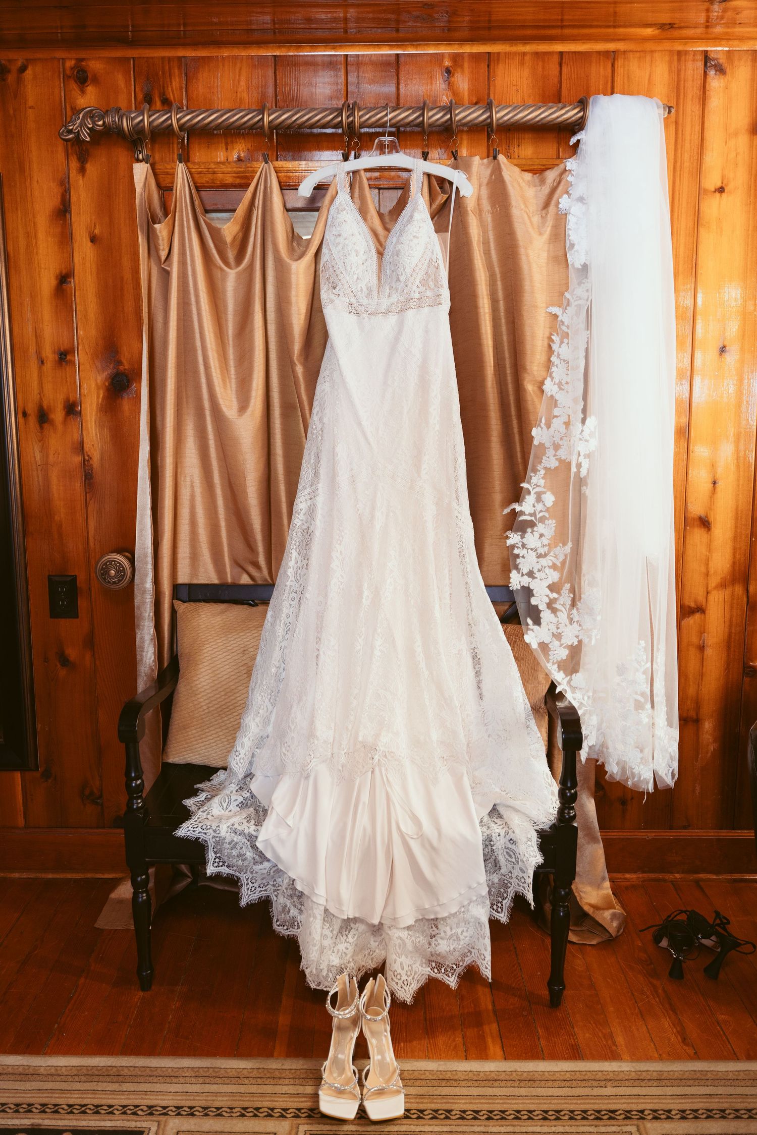 A white lace wedding dress hangs on display with tan bridesmaid dresses and a veil against a wooden wall.