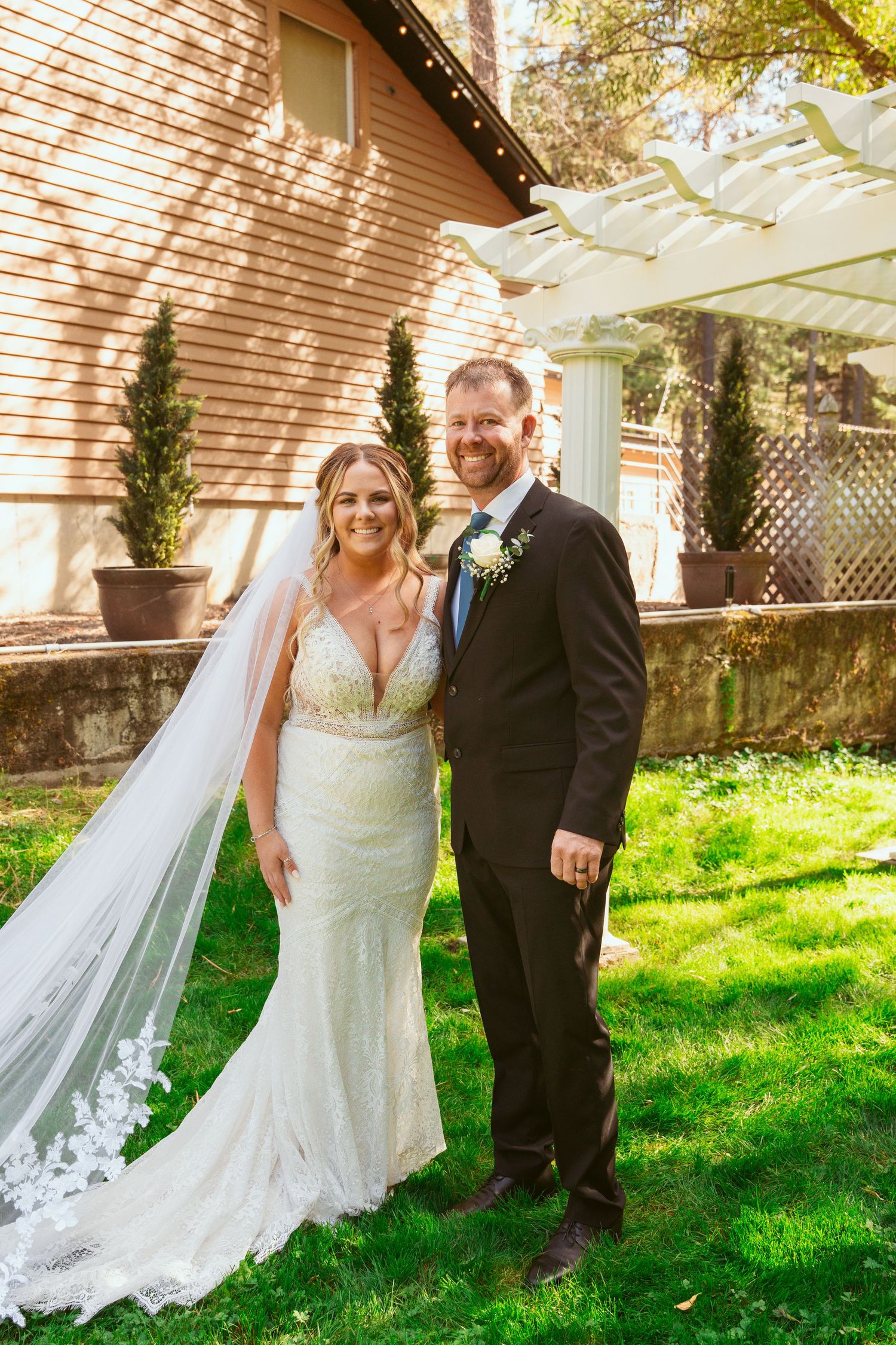 A newlywed couple stands together on grass with a long flowing veil and white pergola in background.