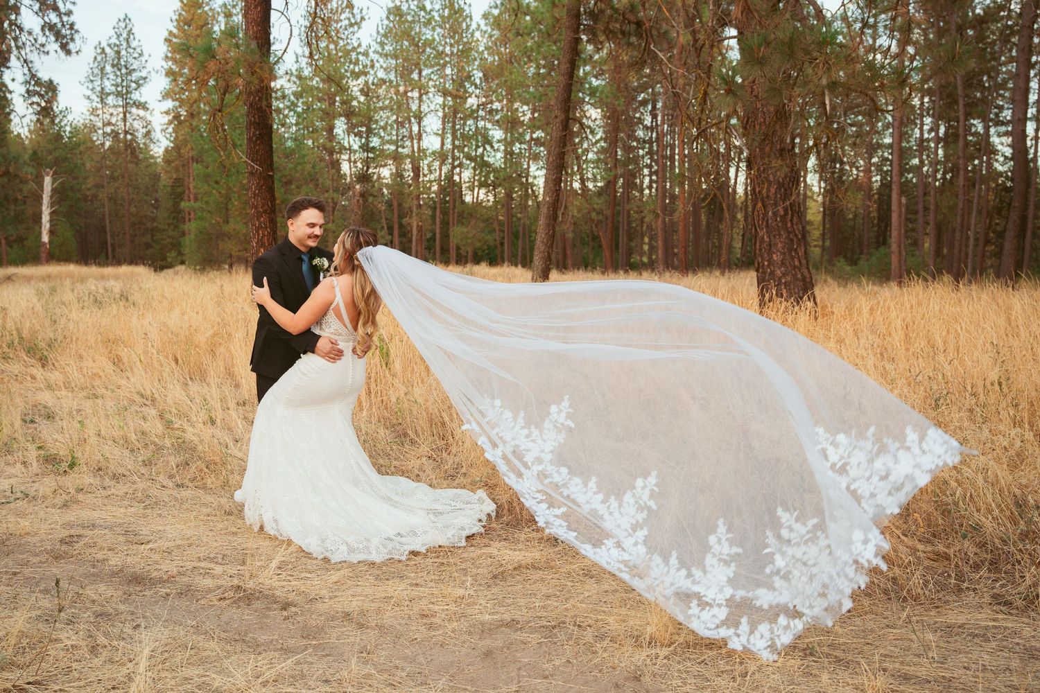 A wedding couple poses with a flowing veil in a scenic pine forest setting.