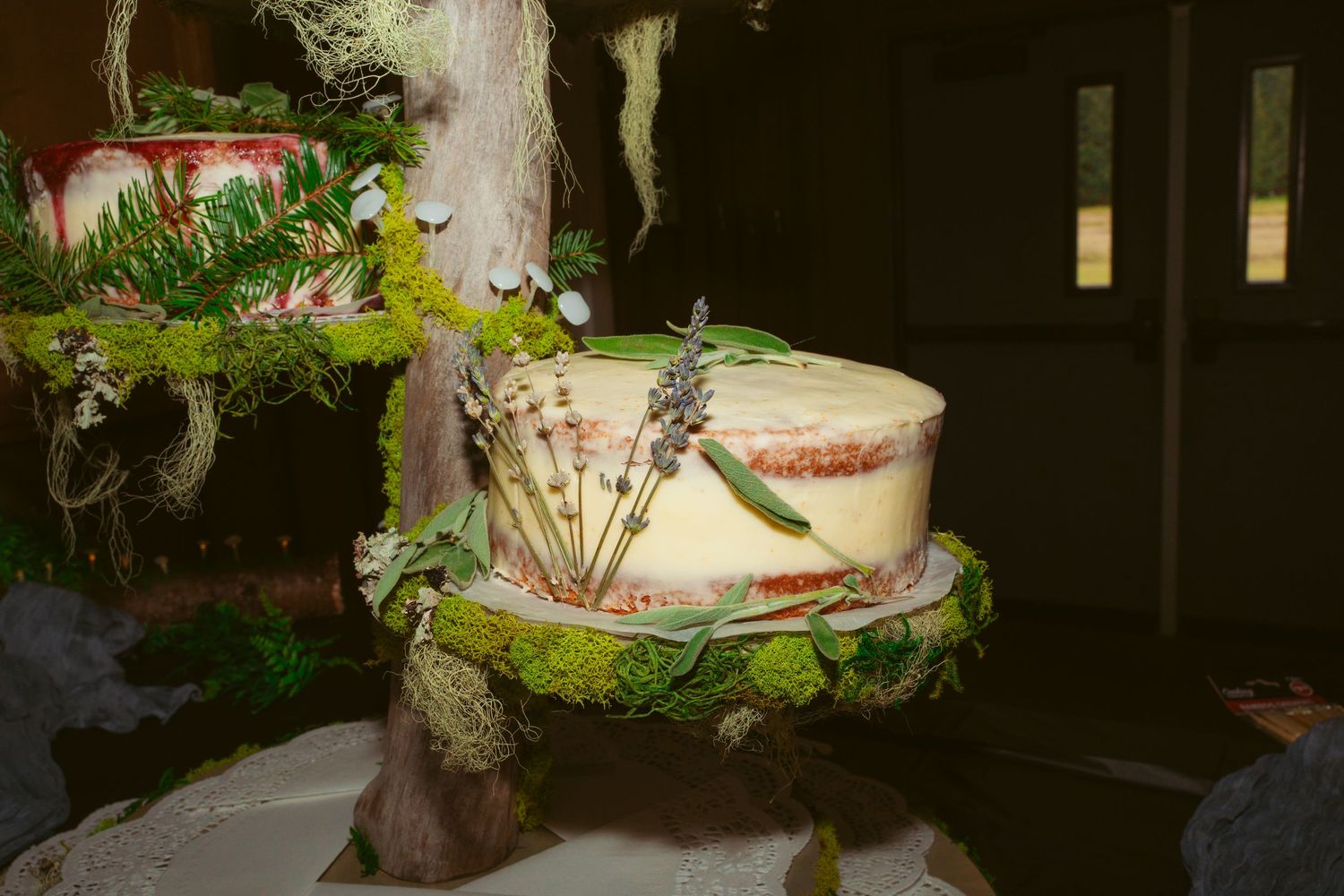 Wedding cake display with natural greenery and rustic decorative elements in dim lighting.
