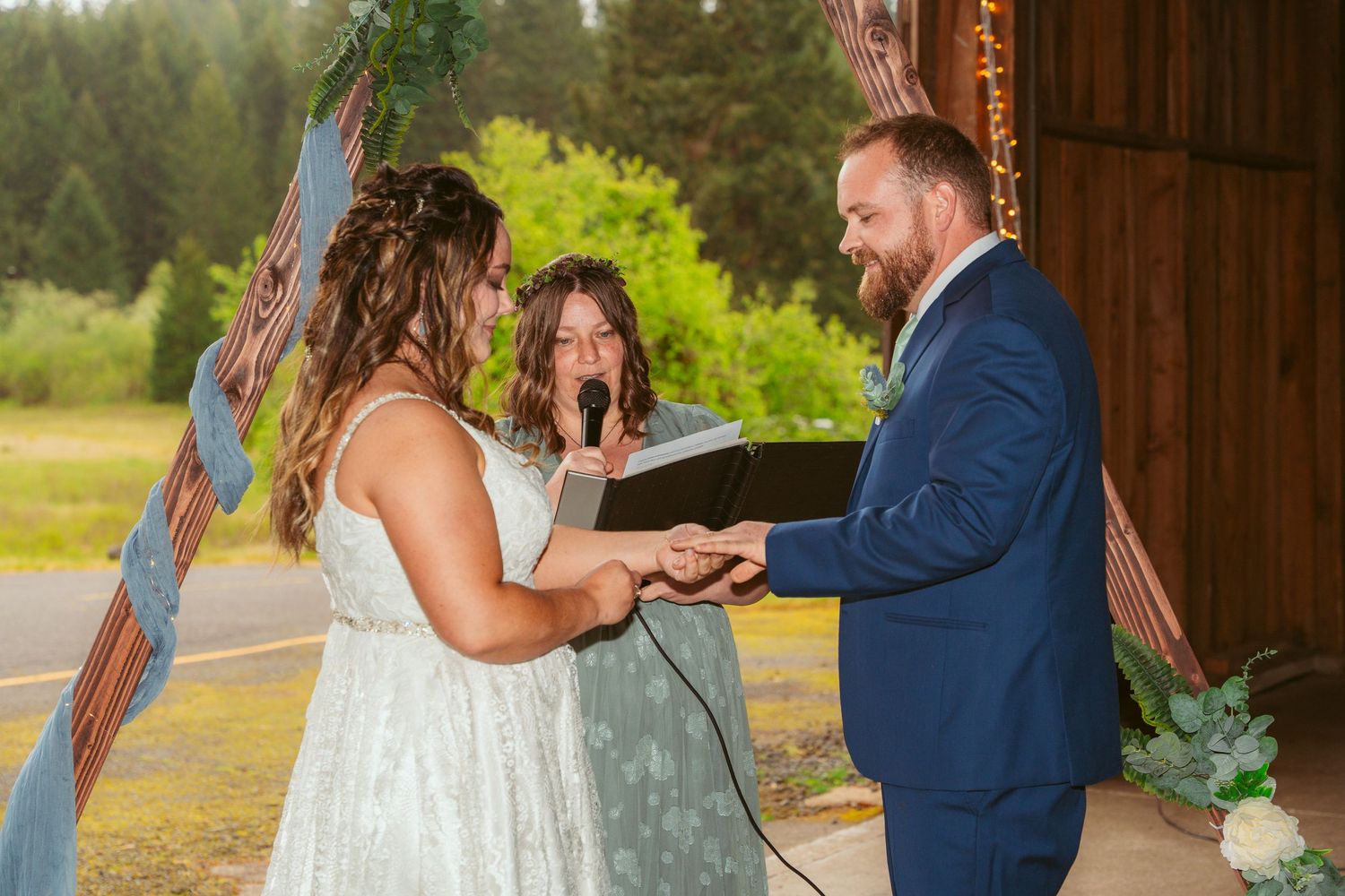 Wedding ceremony exchange taking place in a barn with forest backdrop.