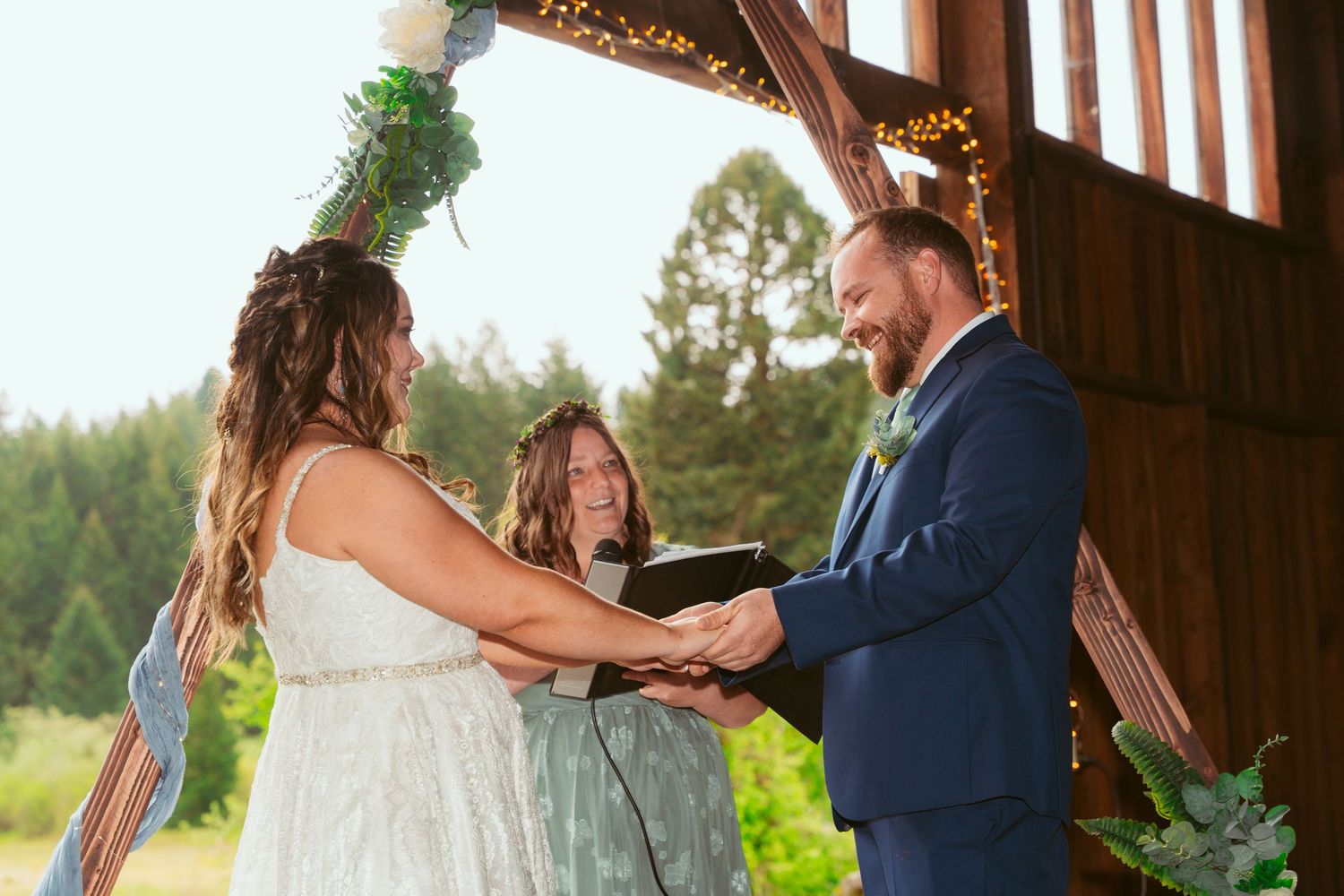 Wedding ceremony taking place in sunlit rustic barn with wooden beams and decorative lighting.